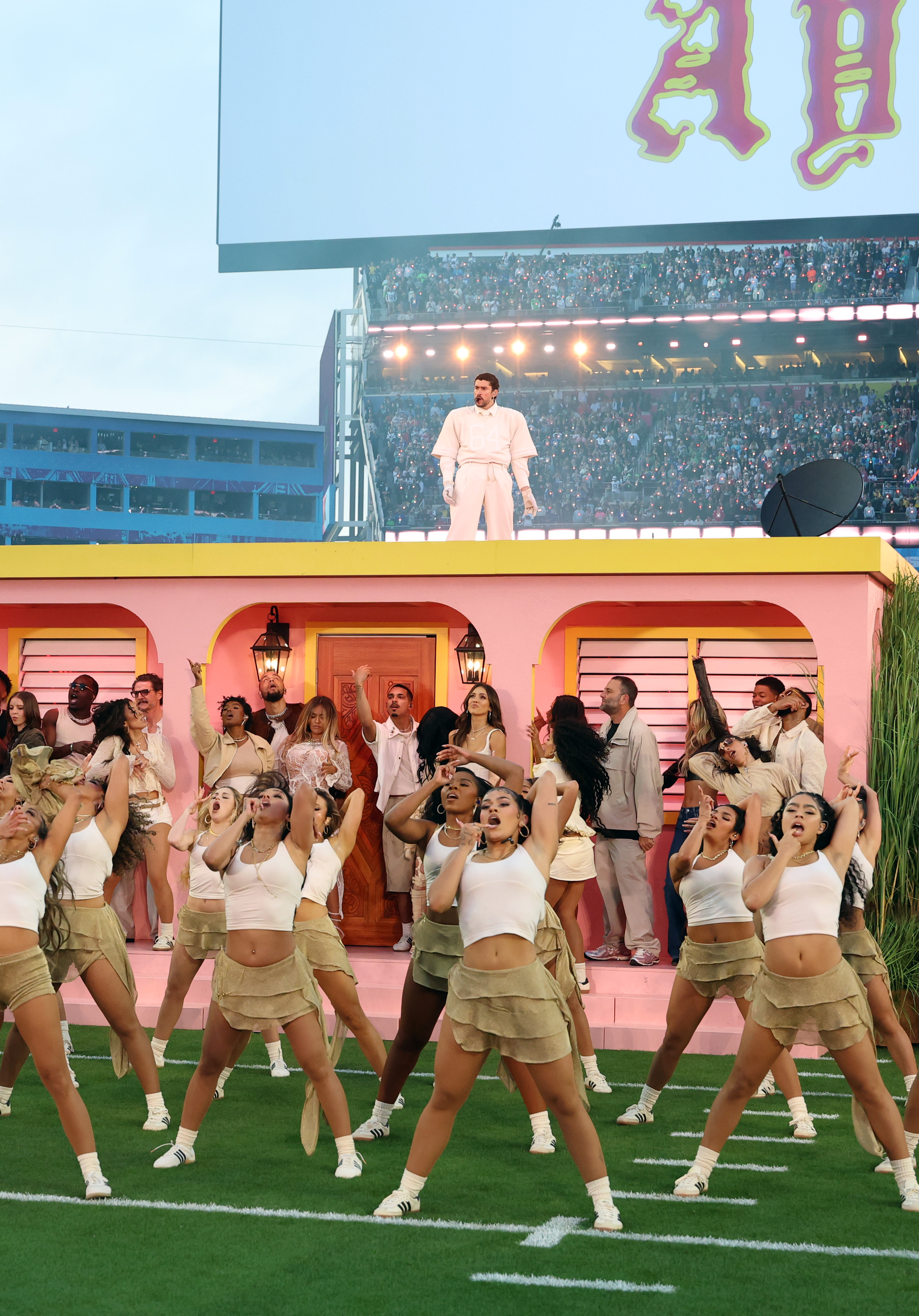 A large group of dancers fills the foreground with synchronized choreography as Bad Bunny stands alone on the roof in an all-cream look, facing the crowd during the 2026 Super Bowl halftime show in Santa Clara, California. | Source: Getty Images