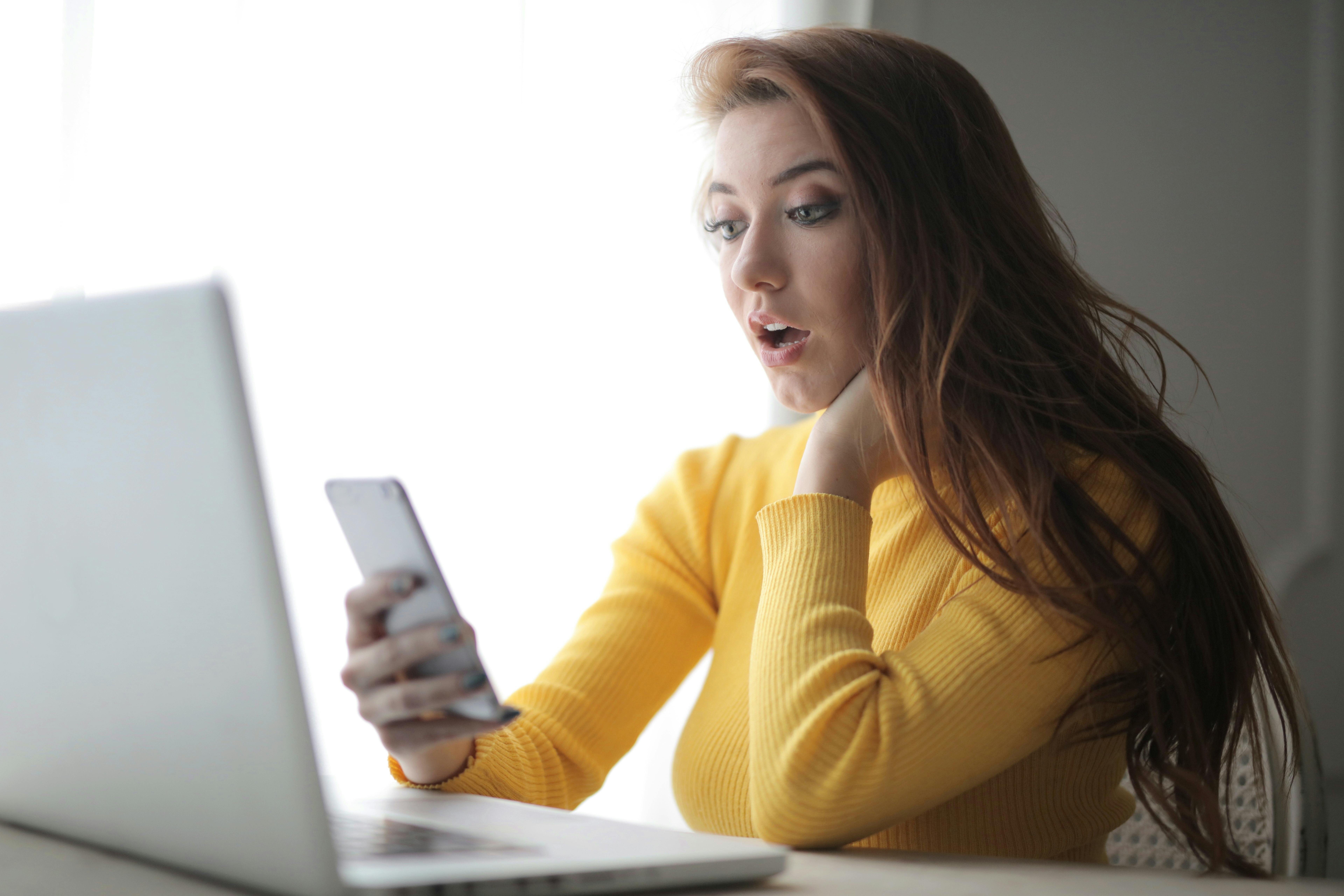 Una mujer sorprendida mirando su teléfono con una computadora portátil sobre un escritorio | Fuente: Pexels