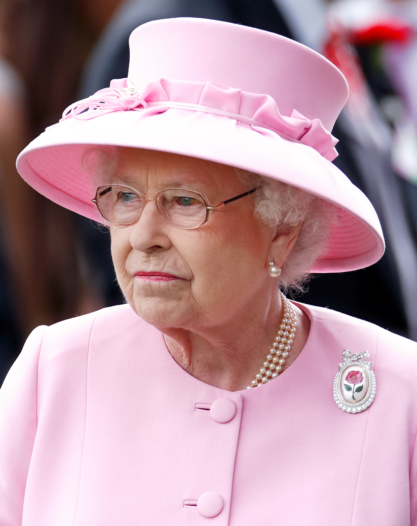 Queen Elizabeth II, seen with a burst blood vessel in her left eye, wears the Centenary Rose Brooch as she attends Day 2 of the 2012 Royal Ascot. Dressed in a pastel pink ensemble and matching hat, the late monarch stands composed during one of her appearances at the prestigious event.