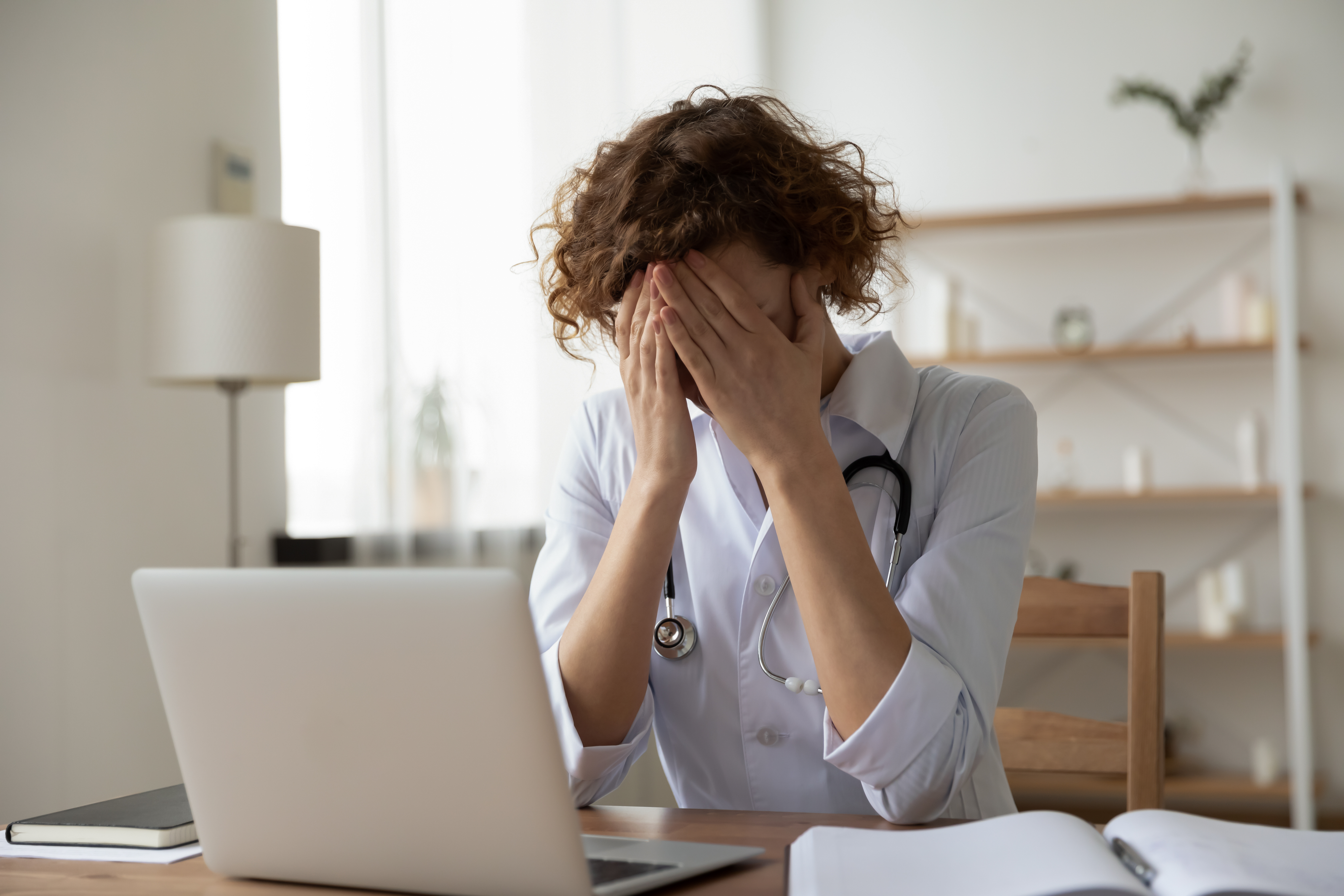 A devastated nurse sitting with her hand over her face while seated in front of a laptop | Source: Shutterstock