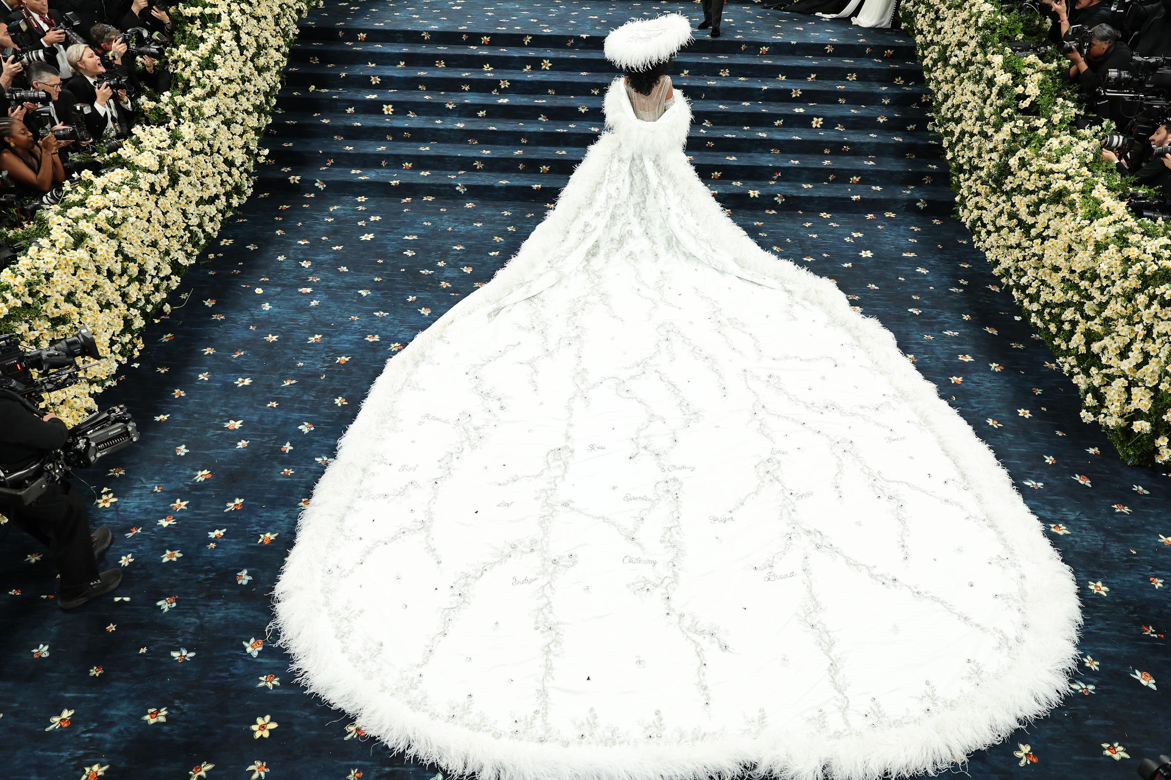 From an aerial angle, the shot captures the grandeur of Ross’s look without showing her face. Her wide-brimmed hat creates a striking focal point, while the full span of her embroidered train unfurls behind her like a work of art.