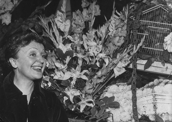Edith Piaf admirant les nombreuses fleurs reçues d'amis, ainsi qu'un cadeau pour une cage à oiseaux, lors d'une réception après sa tournée en Amérique chez Laurent, Paris, le 25 septembre 1957. | Photo : Getty Images