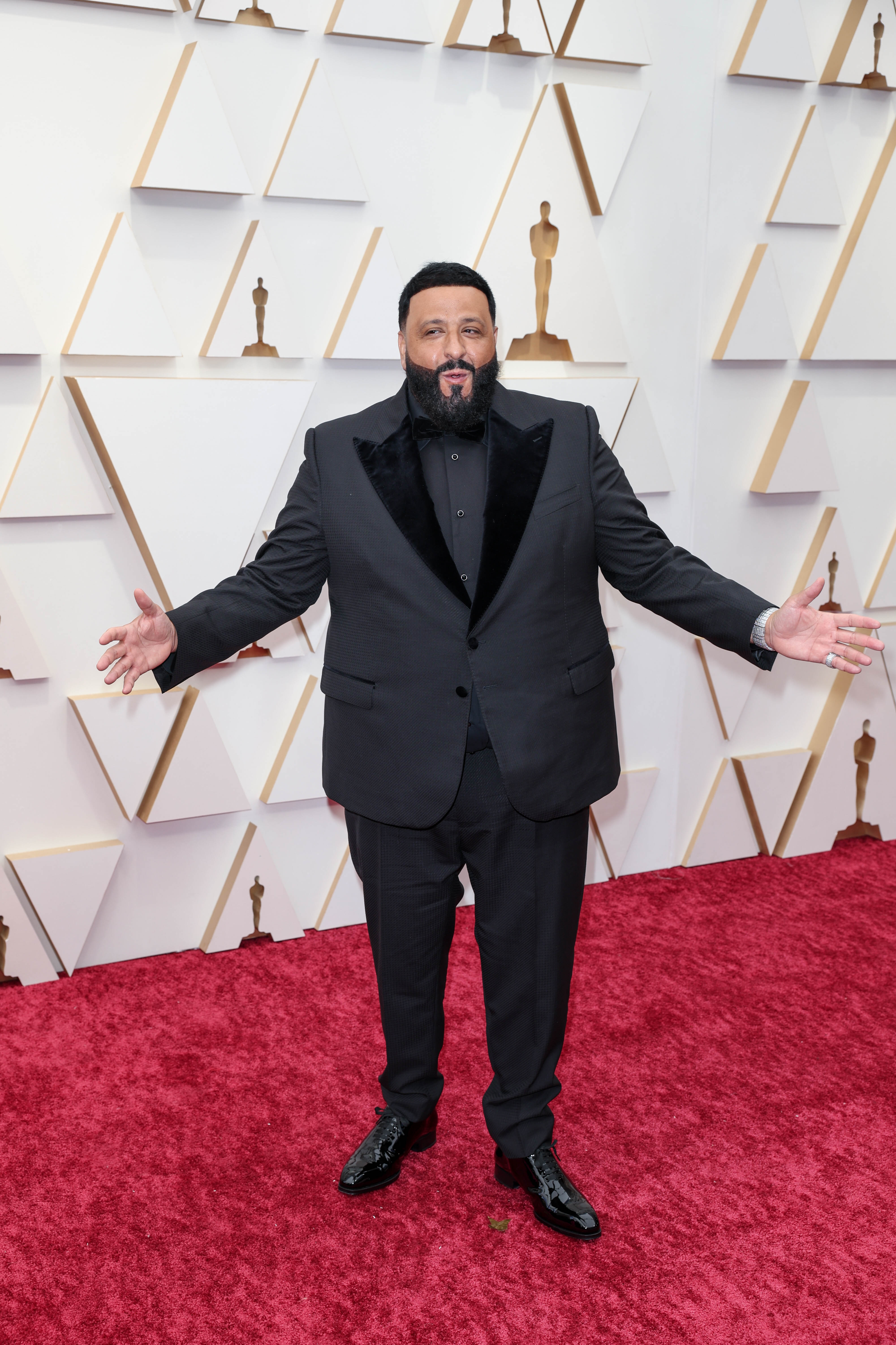 On the Oscars red carpet, DJ Khaled poses in a classic black tuxedo with velvet lapels, arms spread wide against a geometric backdrop adorned with golden statuette silhouettes.