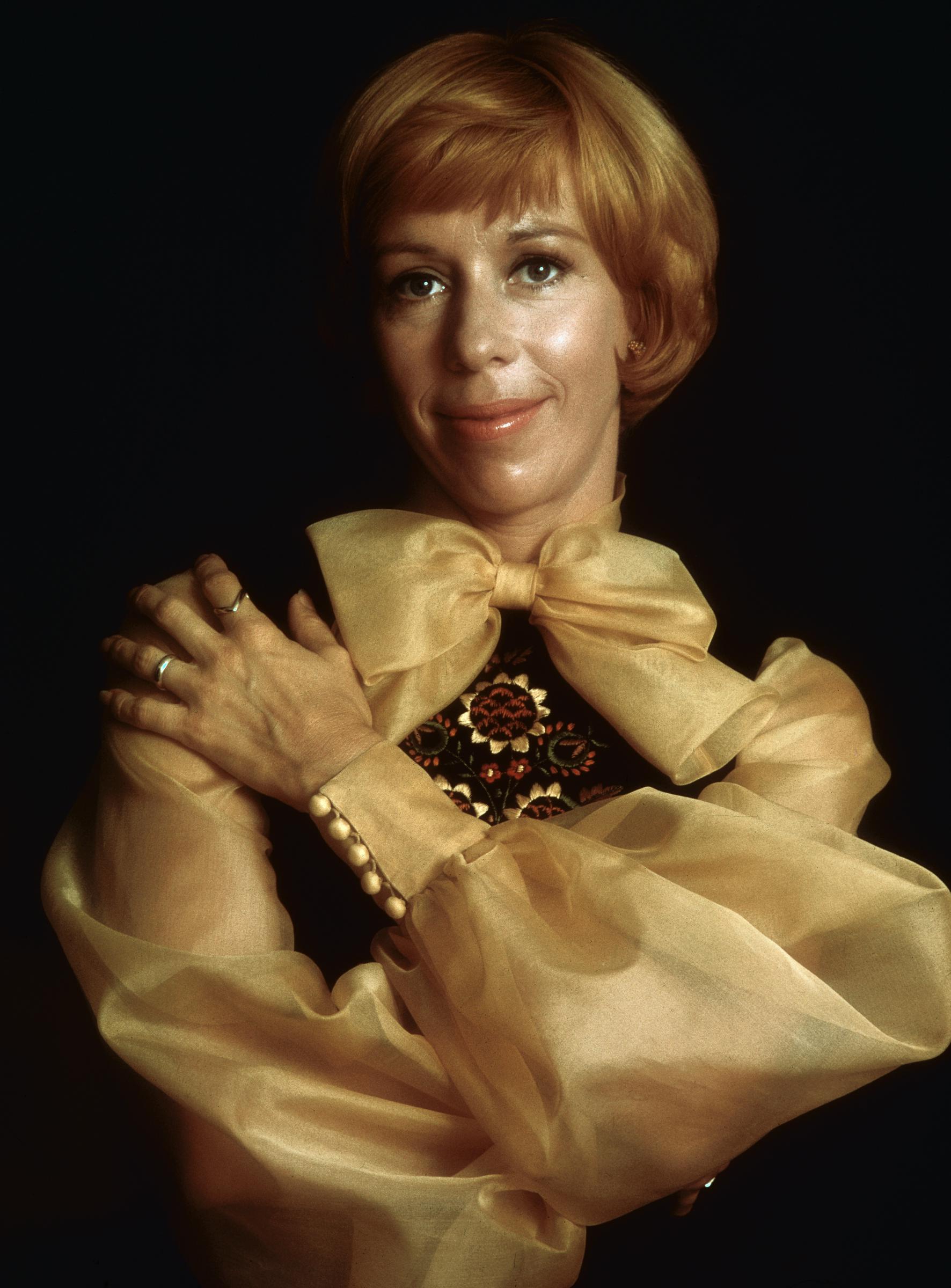 Captured from the waist up, she wears a sheer blouse with voluminous sleeves and a large bow at the neckline over a darker embroidered bodice. Her arms are crossed lightly over her chest as she smiles toward the camera.