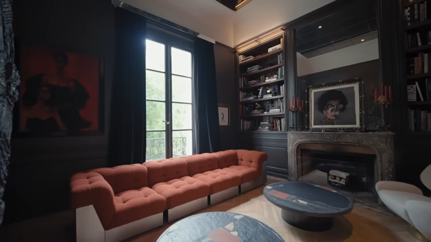 The warm, sophisticated library featuring dark espresso-painted wood panels, custom bookshelves, a stone fireplace, and a plush rust-colored modular sofa. | Source: YouTube/@Archdigest