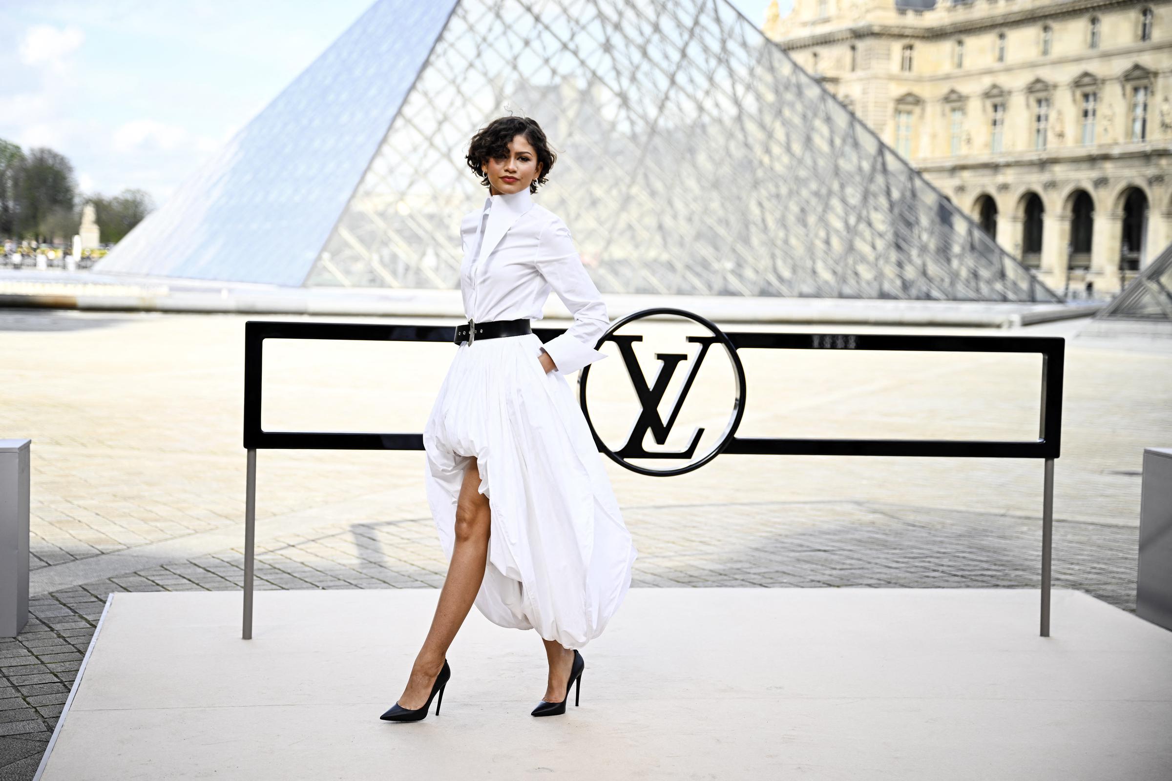 Zendaya poses beside a black railing featuring the Louis Vuitton “LV” logo with the Louvre’s glass pyramid behind her during the Louis Vuitton Fall/Winter show at Paris Fashion Week in Paris on March 10, 2026. | Source: Getty Images