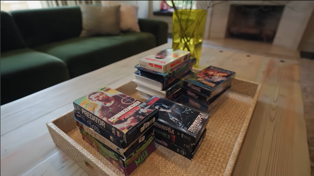 A woven tray on the light wood coffee table displays stacks of classic VHS tapes, from "Predator" to "Ghost." | Source: YouTube/@Archdigest