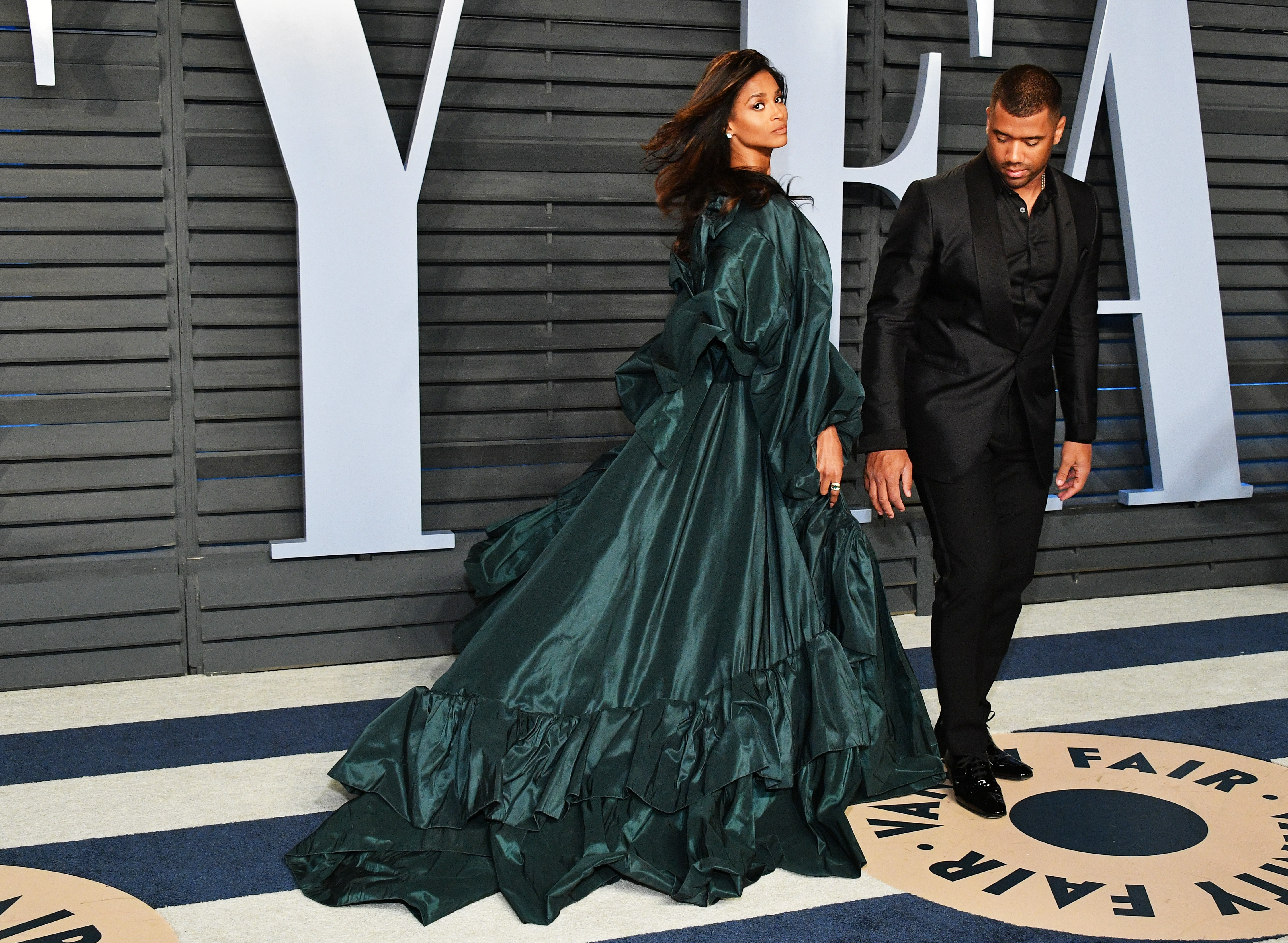 Ciara turns mid-step in a dramatic flowing green gown, the fabric sweeping around her as she moves with striking confidence. Russell Wilson walks just behind her in a tailored black suit, keeping a steady, composed stride that contrasts her motion.