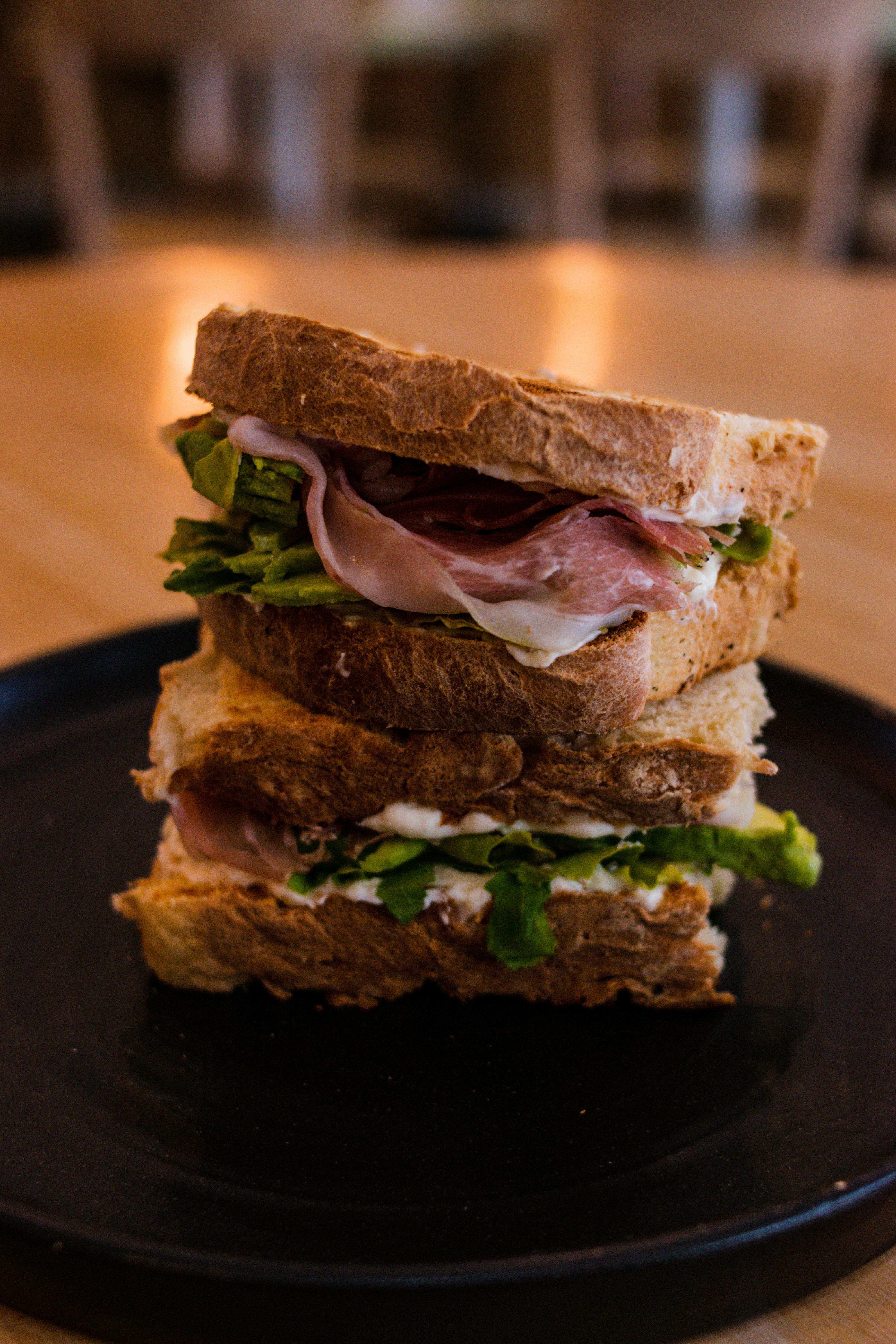 Stacked wholegrain sandwich layered with cured meat, greens, and creamy spread, presented on a dark plate in a warm, softly lit setting. | Source: Pexels