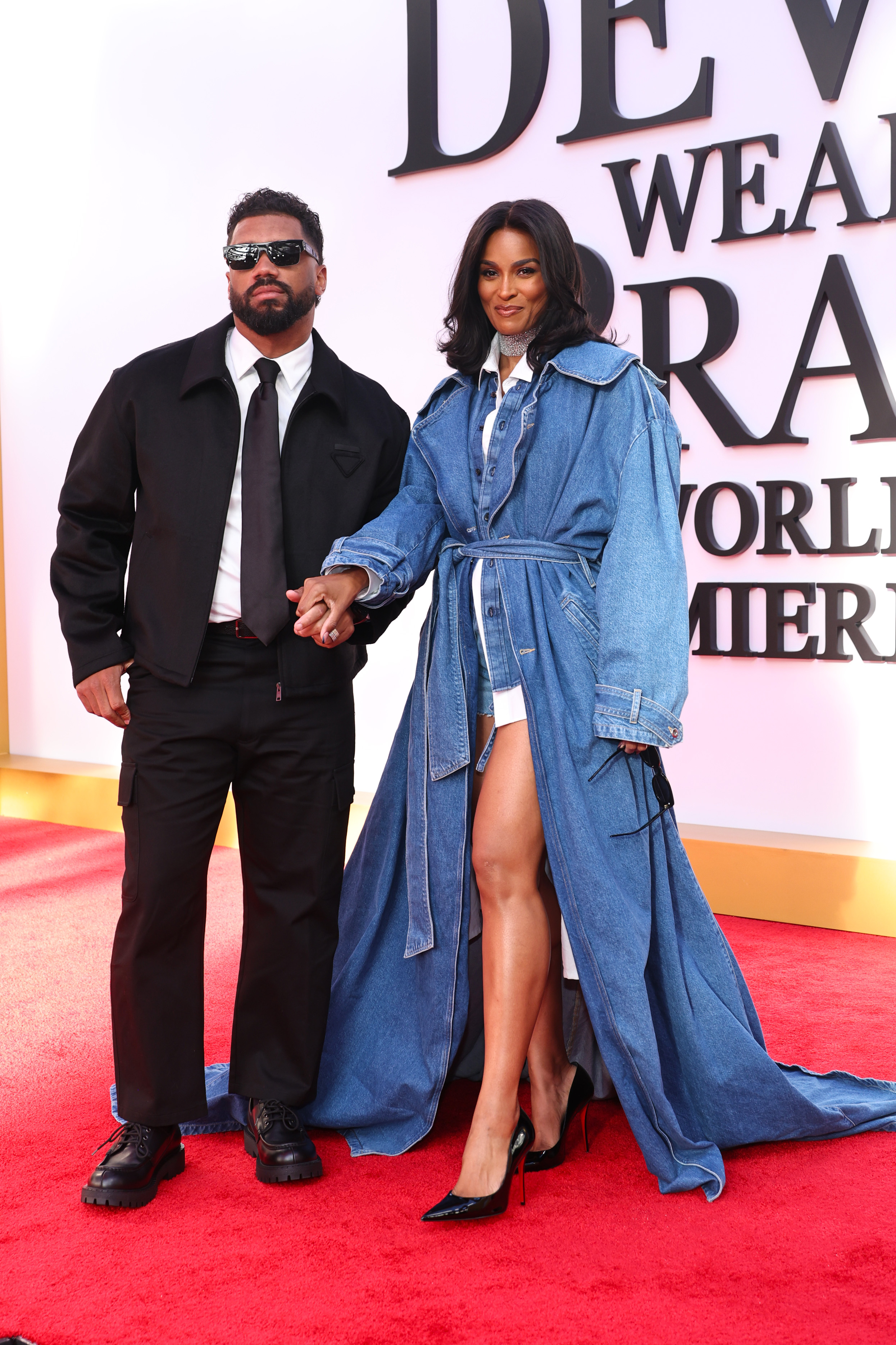Russell Wilson and Ciara pause for photos on the carpet, holding hands as they face forward with calm, confident expressions. Her high-slit look and statement accessories bring a bold finish, while his all-black ensemble grounds the moment.