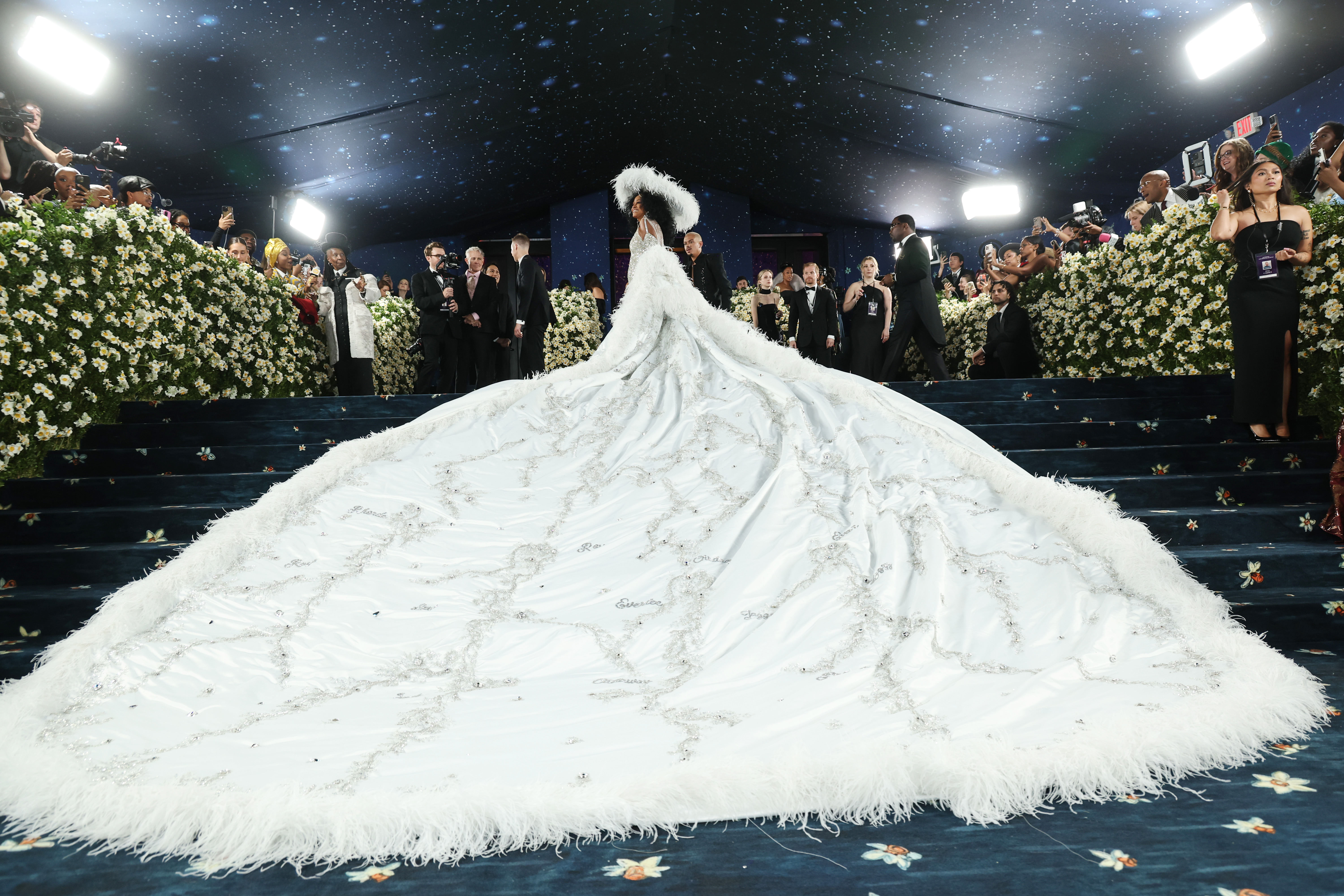 Captured from the base of the stairs, this breathtaking shot frames Ross at the very top, like a vision in motion. The angle showcases the full splendor of her gown, from the intricate embroidery and delicate feather-like textures to the sheer magnitude of the 18-foot train cascading behind her.
