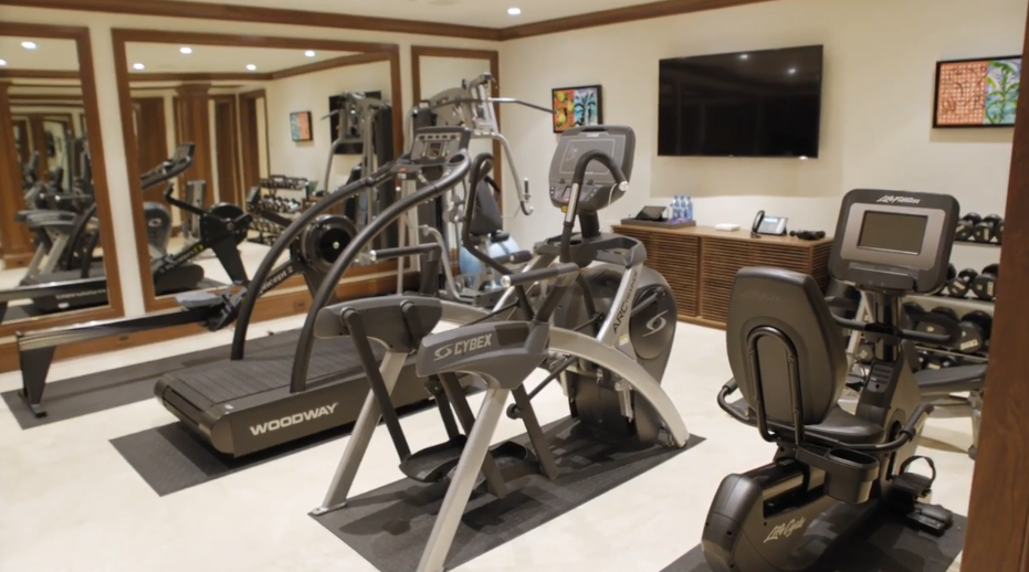 Equipped with state-of-the-art exercise machinery, the home gym includes treadmills, elliptical trainers, stationary bikes, and weight racks mirrored along the wall. A flat-screen television and soft cream walls give the workout space a sleek, professional aesthetic. | Source: YouTube/Architectural Digest