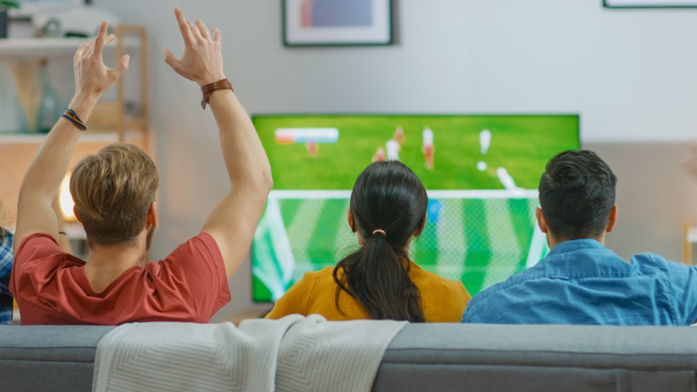 En casa, varios hinchas deportivos sentados en un sofá viendo un importante partido de fútbol en la televisión. I Foto: Shutterstock En casa, varios hinchas deportivos sentados en un sofá viendo un importante partido de fútbol en la televisión. I Foto: Shutterstock