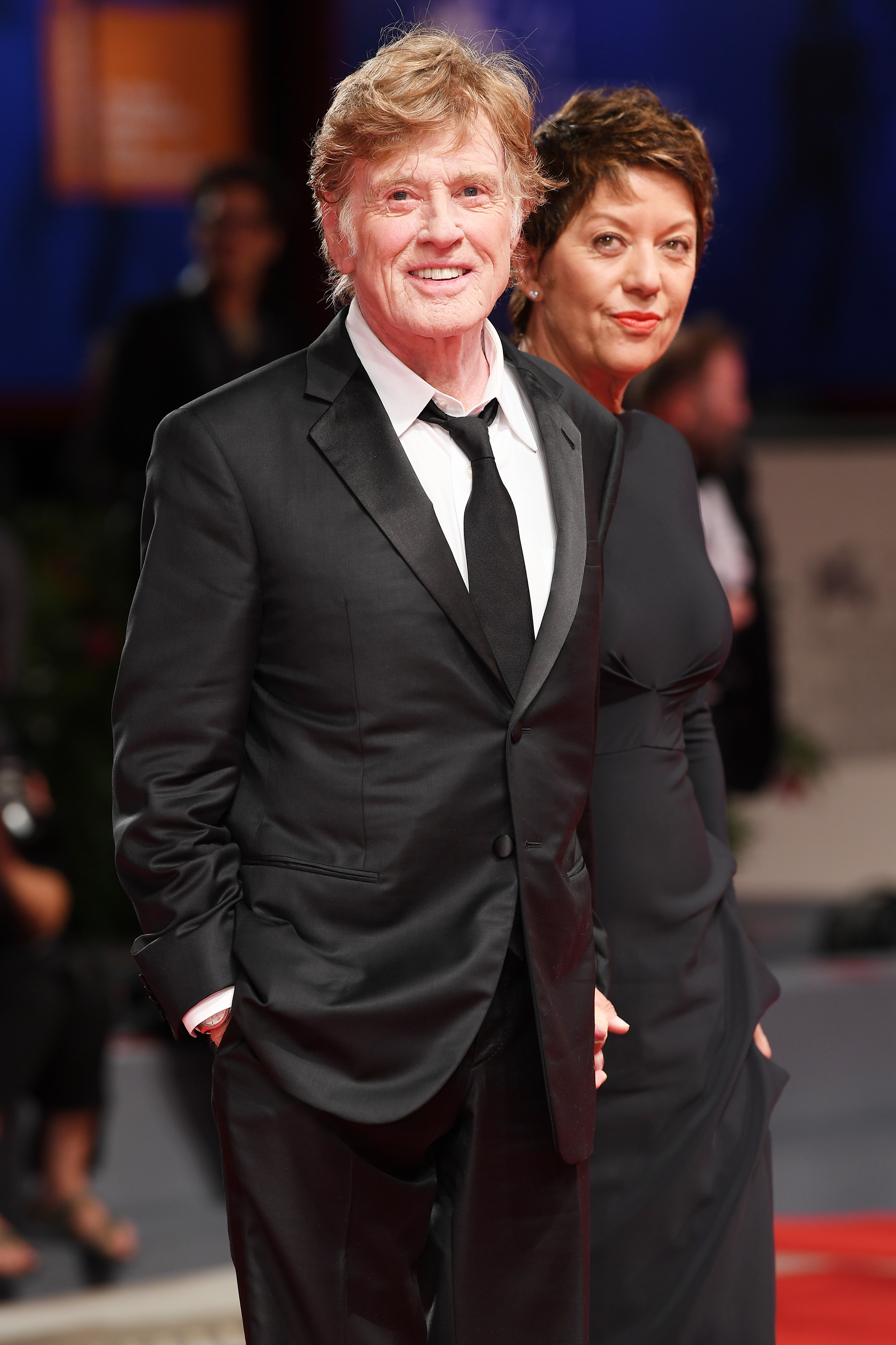 Robert Redford and Sibylle Szaggars walk the red carpet for the "Our Souls at Night" screening at the Venice Film Festival on September 1. Szaggars wears a black gown with a fitted bodice and draped detailing. Redford wears a black suit, white shirt, and black tie. They are pictured in front of the press line.