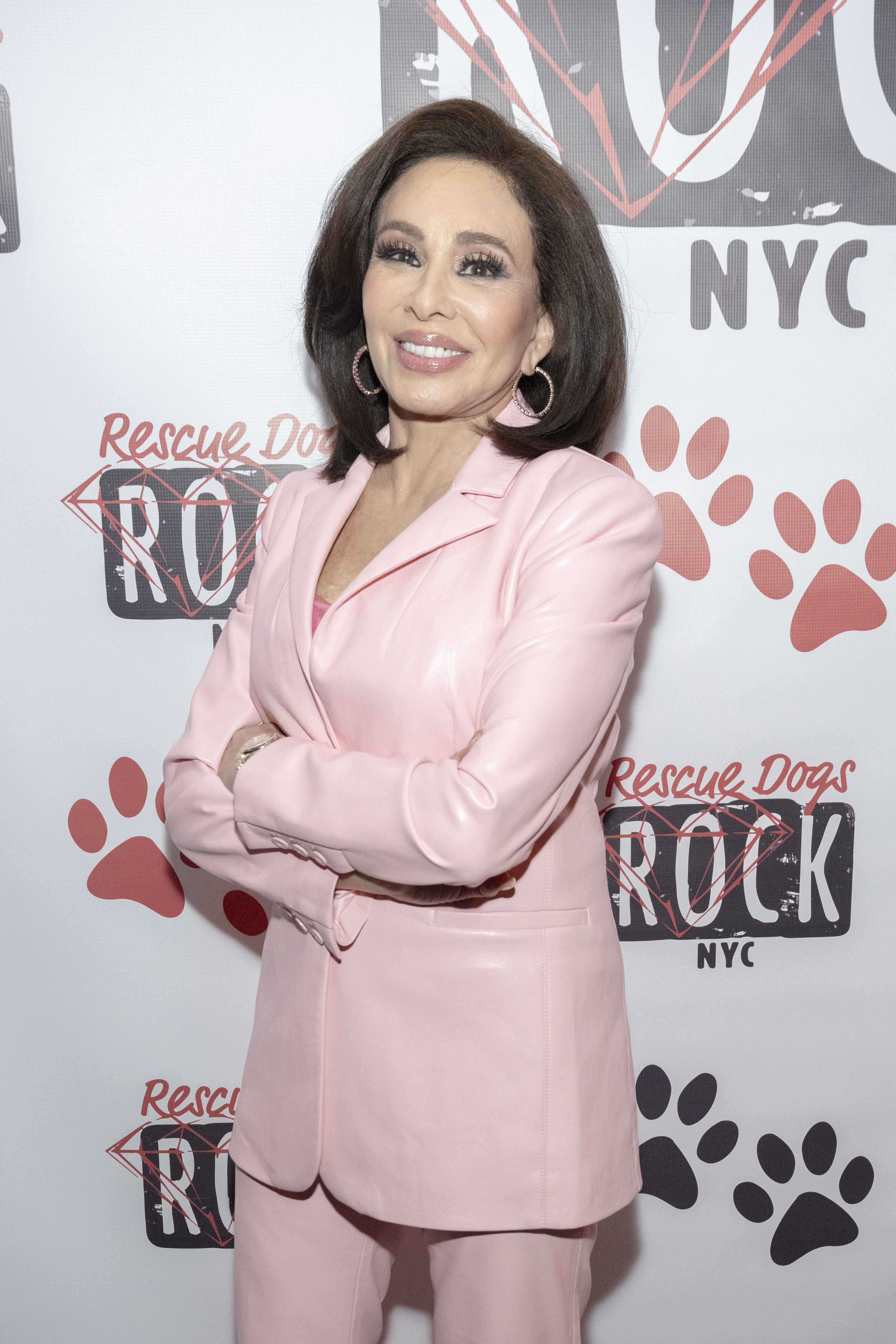 Jeanine Ferris Pirro attends the Rescue Dogs Rock NYC charity cocktail event, donning a pastel pink tailored suit with her arms folded. Her expression is warm and composed, fitting the advocacy‑focused gathering.