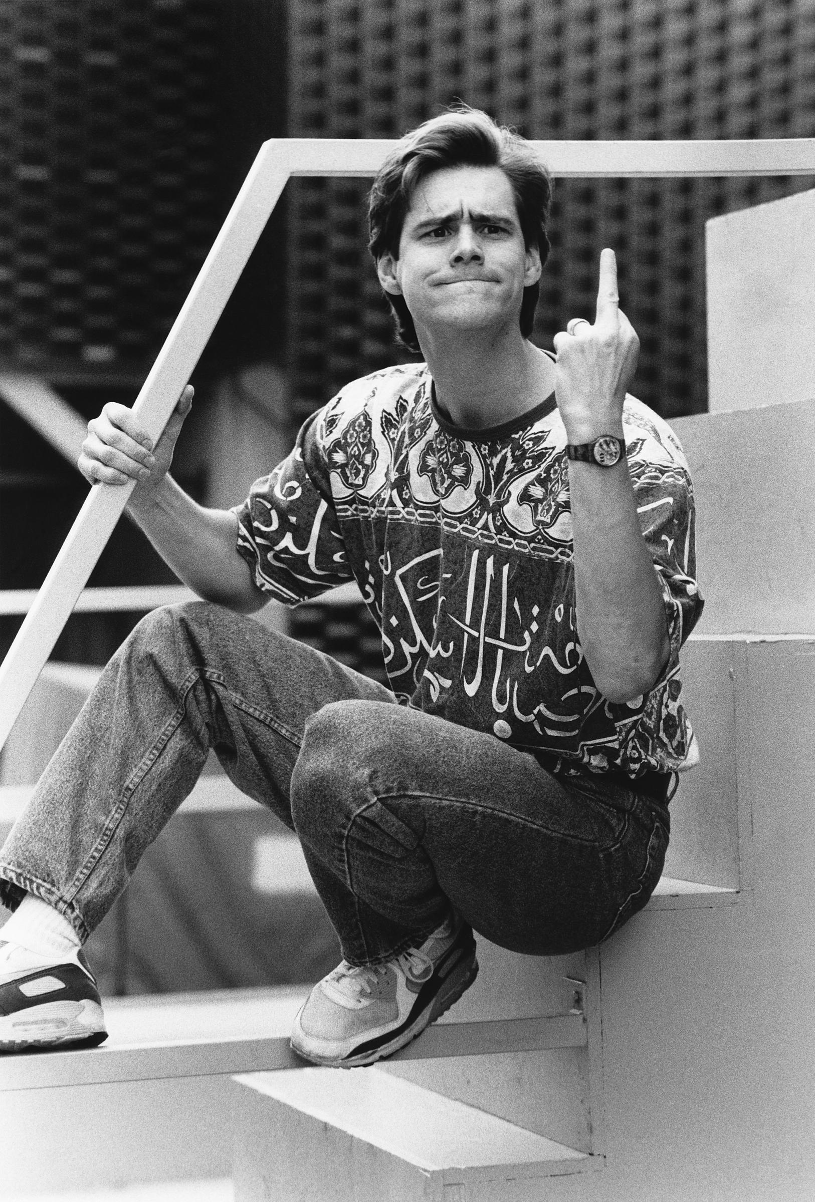 Jim Carrey is seated casually on modern outdoor steps, wearing patterned casual clothes and sneakers in this black-and-white photo. With a playful yet defiant expression, he raises his finger toward the camera in classic rebellious humor. The image captures his trademark blend of comedy, confidence, and irreverent charm.