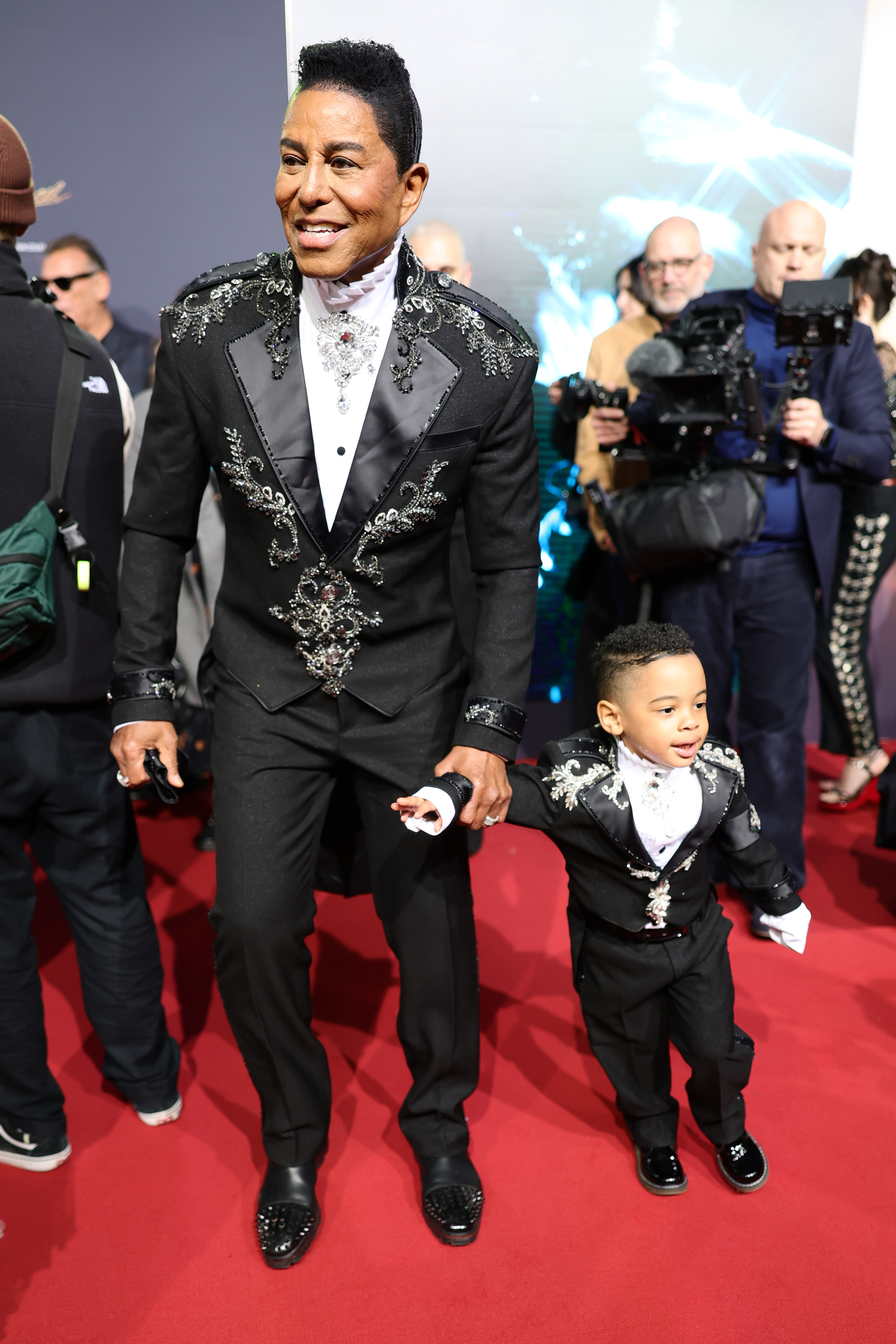 Jermaine Jackson walks hand-in-hand with his son at the premiere, their matching embellished suits creating a standout father-son moment. The child’s miniature version—complete with crisp white shirt and polished shoes — mirrors his father’s look, adding a charming, eye-catching symmetry.
