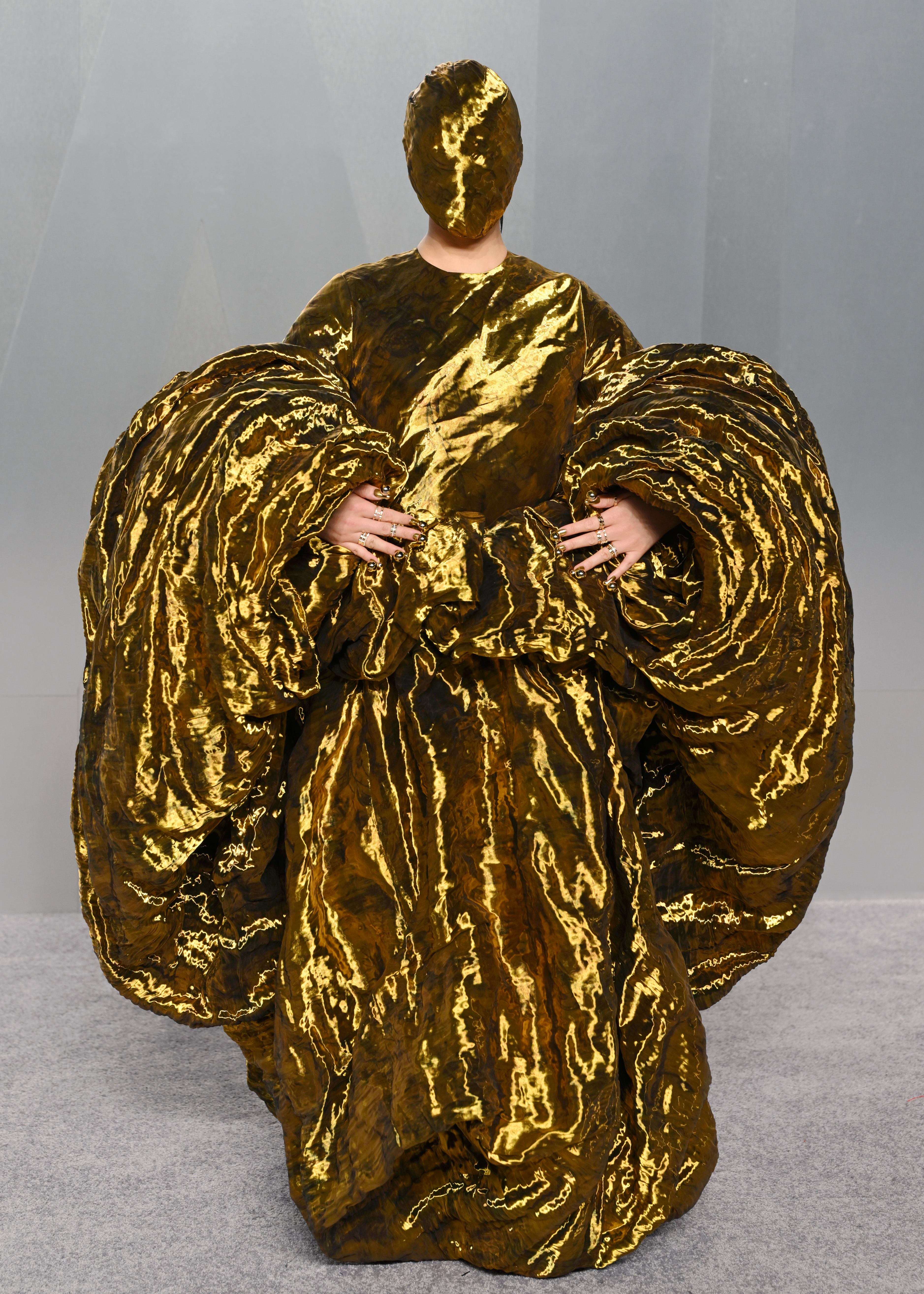 Audrey Nuna is completely unrecognisable at the Vanity Fair Oscar Party, her entire figure enveloped in a voluminous floor-length gold metallic sculptural gown with enormous swirling coiled sleeves; only her bejewelled hands are visible, her face entirely concealed behind a smooth, featureless gold mask.