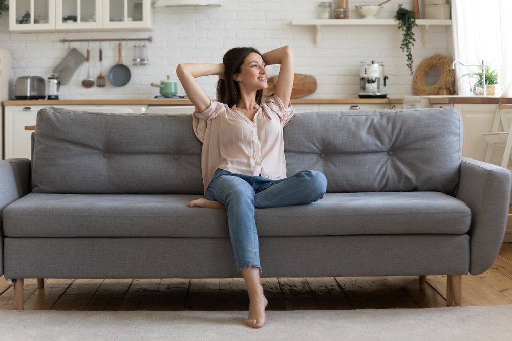 A relieved and happy young woman sitting in a cozy living room with her hands behind her head | Photo: Shutterstock/fizkes