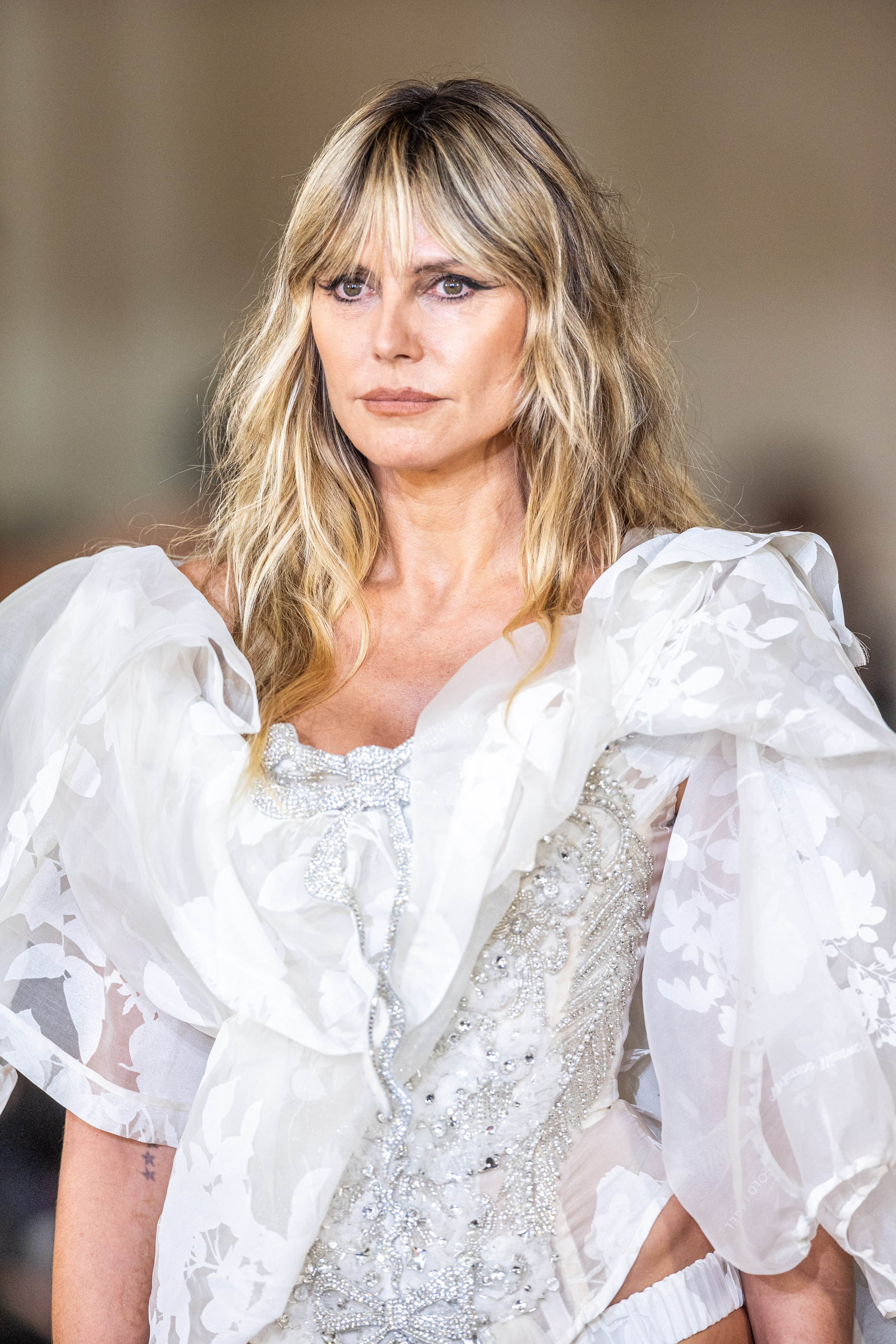 Heidi Klum walked the runway at the Vivienne Westwood Ready-to-Wear Spring/Summer 2026 show during Paris Fashion Week in France, wearing a white dress. The bodice was adorned with silver filigree and featured an ornate, bejeweled bow on the plunging neckline. Sheer white embroidered fabric added puffy sleeves and a cape-like detail, enhancing the dramatic elegance of her look.