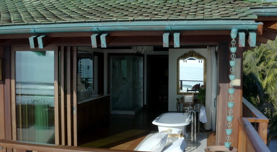 An exterior view into the upper-level suite reveals folding glass doors that open the entire bathroom to the sea breeze. A glass-enclosed shower, a double vanity, and a clawfoot tub are framed by dark timber posts, a green-tiled roof overhang, and copper rain chains hanging along the balcony edge. | Source: YouTube/Architectural Digest