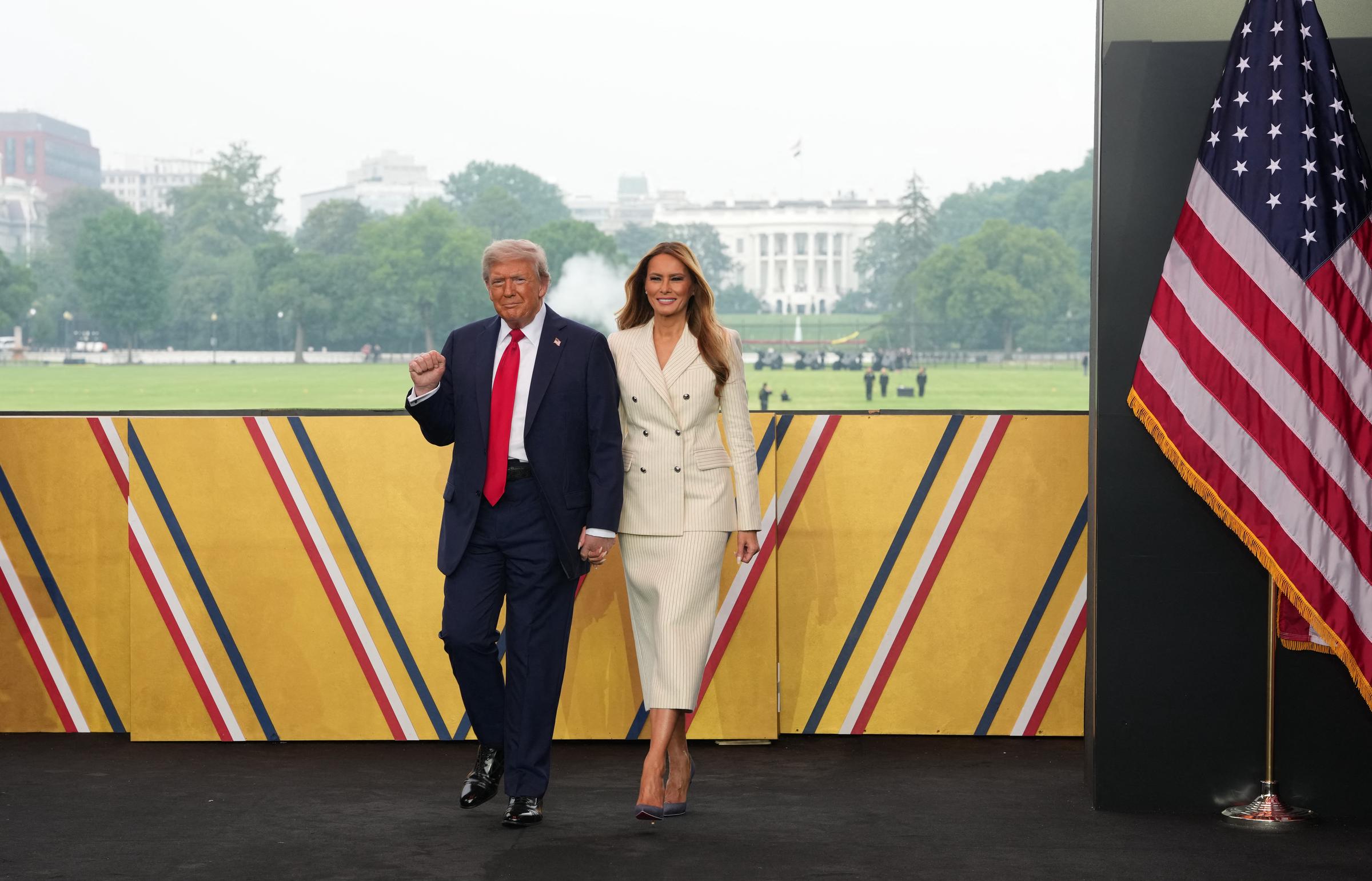 Donald Trump walks alongside Melania Trump, raising his fist slightly as they approach the stage at the Army’s 250th Anniversary Parade on June 14, 2025. The White House stands in the background behind a patriotic backdrop and American flag.
