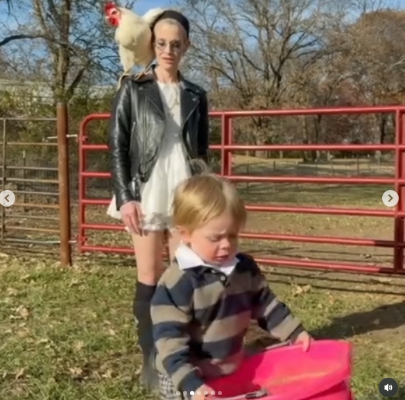 Kelly Osbourne brought her son, Sidney, along for farm chores, with the youngster carrying a bright pink container as his mother followed behind in a leather jacket and boots. A chicken perched on Osbourne's shoulder, capturing the hands-on, animal-filled routine the family has embraced in Iowa. The moment appeared in a post dated November 14, 2025. | Source: Instagram/kellyosbourne