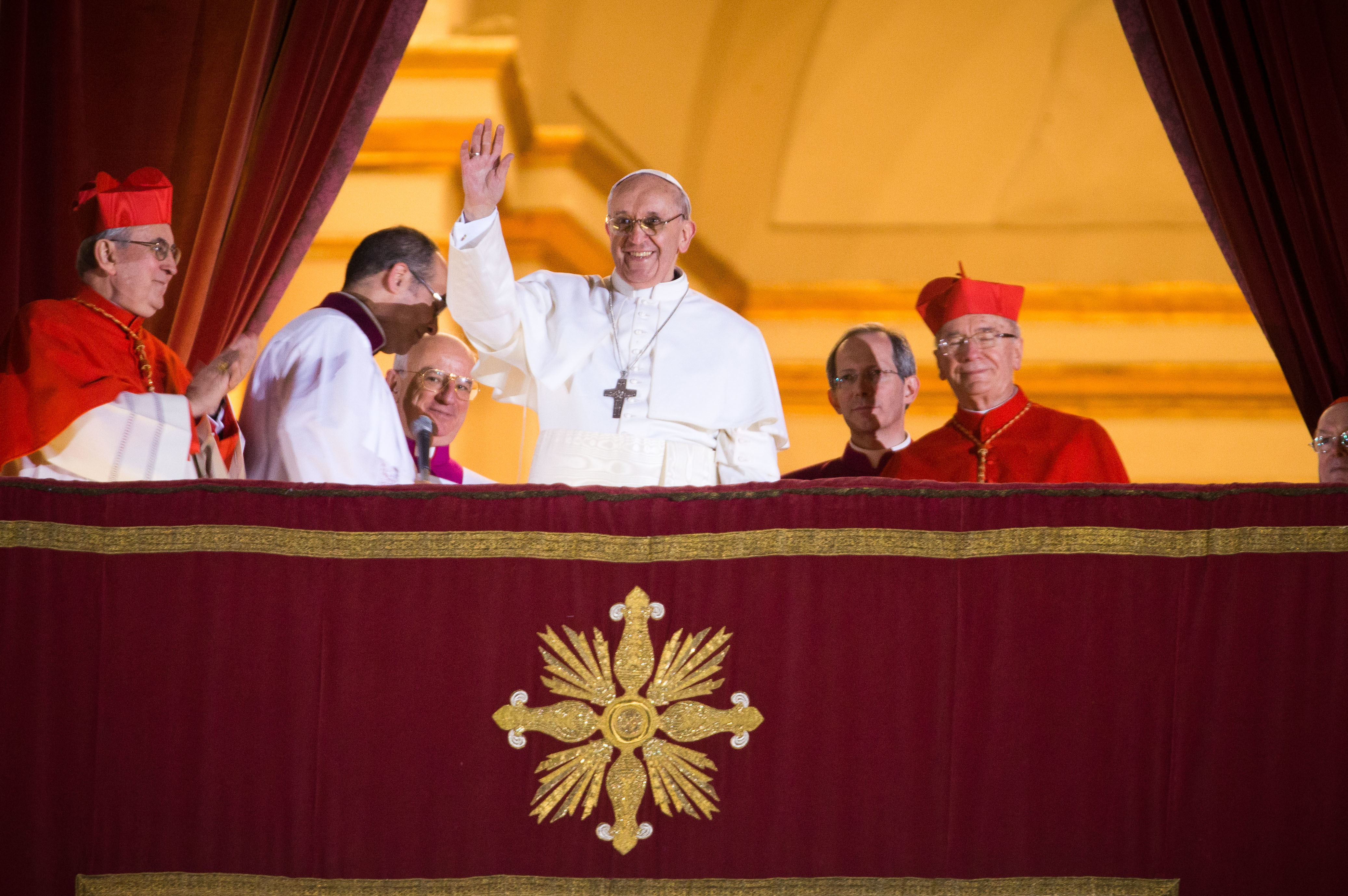 Pope Francis waves from the central balcony, smiling in white robes and silver cross, surrounded by cardinals in scarlet under a golden-lit archway