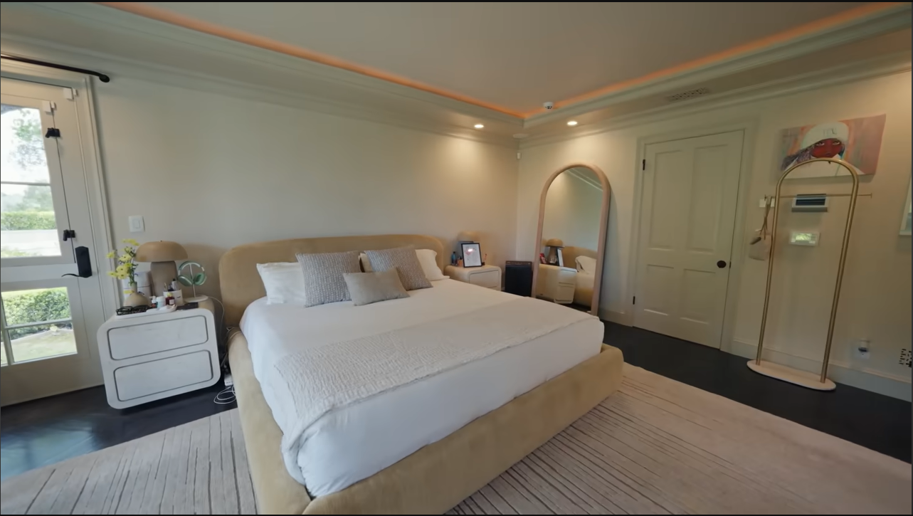 Palmer's cozy, neutral-toned master bedroom features a sprawling bed, warm recessed lighting, and a massive arched floor mirror. | Source: YouTube/@Archdigest