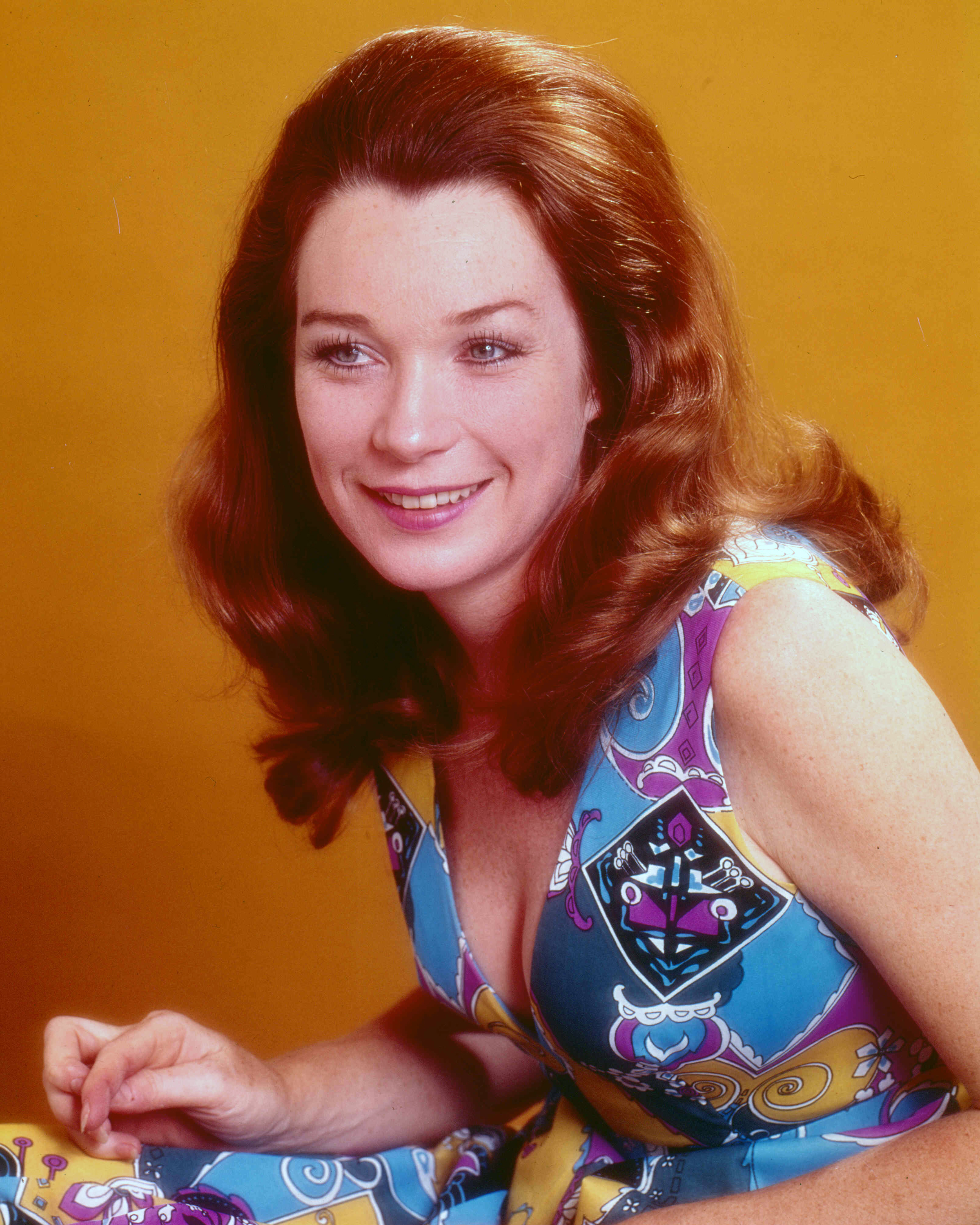 Shirley MacLaine wearing a sleeveless print pattern dress and smiling in a studio portrait against an orange background, circa 1970.