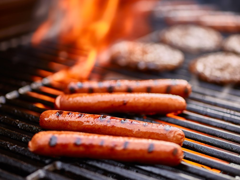 Fot dogs savoureux en train de cuire sur un grill avec des hamburgers. | Shutterstock Fot dogs savoureux en train de cuire sur un grill avec des hamburgers. | Shutterstock