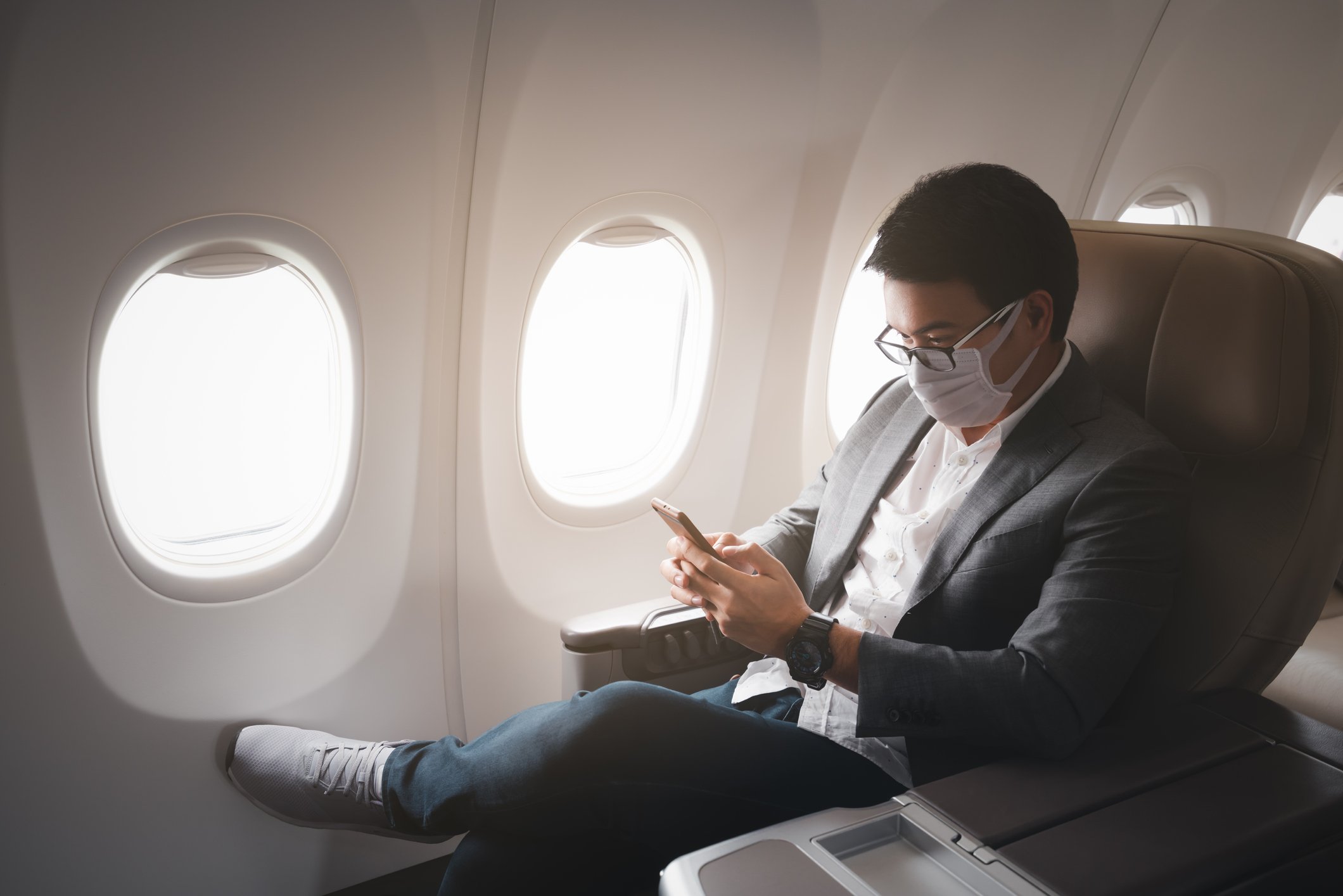 Junger Geschäftsmann sitzt mit aufgesetzter Gesichtsmaske im Flugzeug und benutzt sein Smartphone. | Quelle: Getty Images