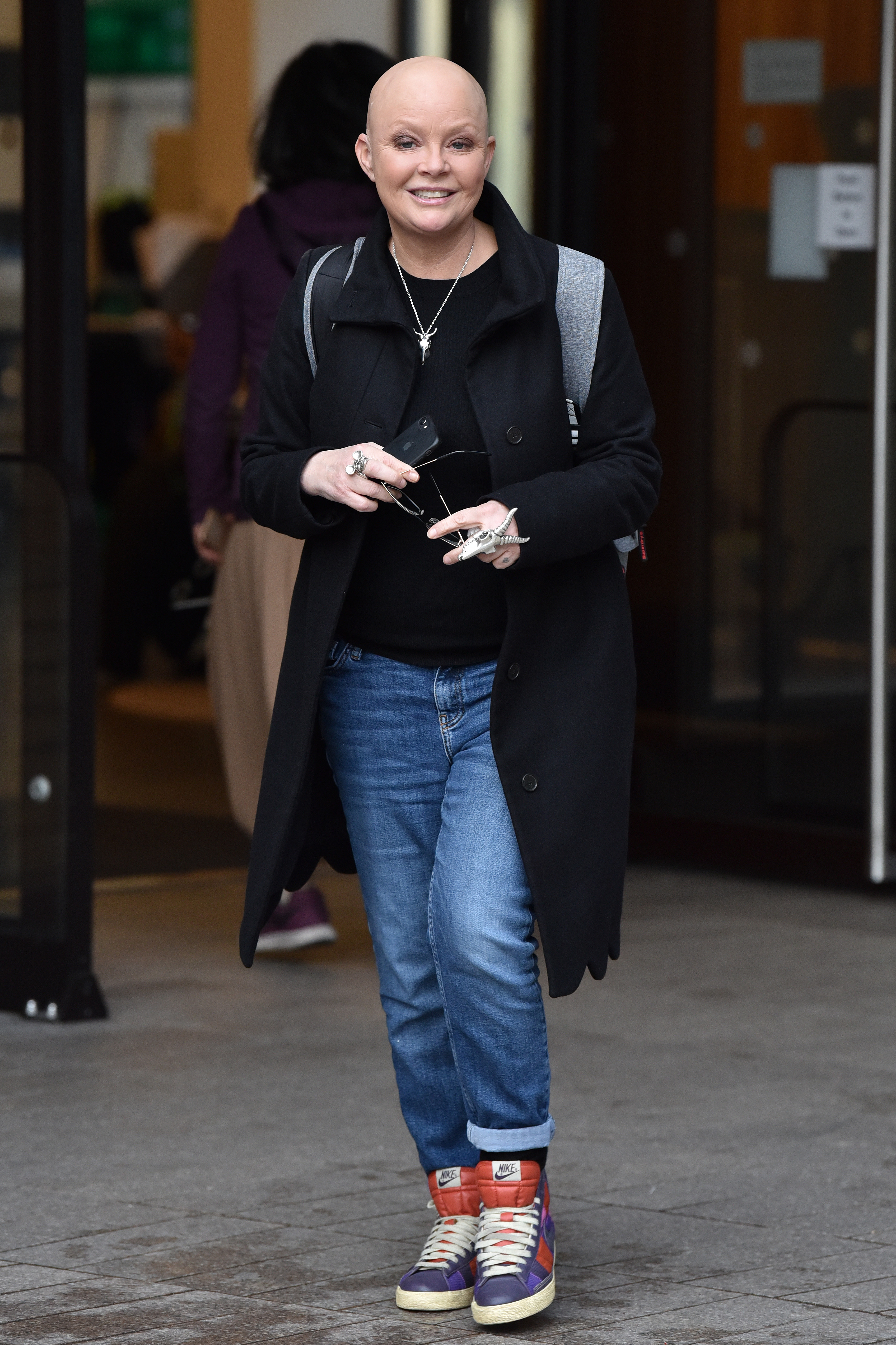 Porter strides out of a building grinning broadly, wearing a long black double-breasted coat over a black top and loose-fit cuffed jeans, finished with chunky multicoloured Nike high-top trainers. She holds a pair of sunglasses in one hand and carries a backpack. Pictured outside ITV Studios, London, January 2020.