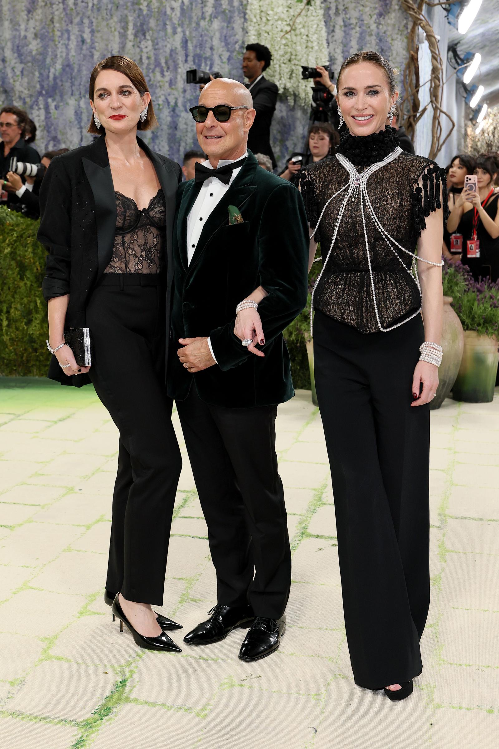 Felicity Blunt and Emily Blunt coordinate in sheer black lace and sharply tailored silhouettes. Stanley Tucci anchors the trio in a velvet tuxedo with dark sunglasses.