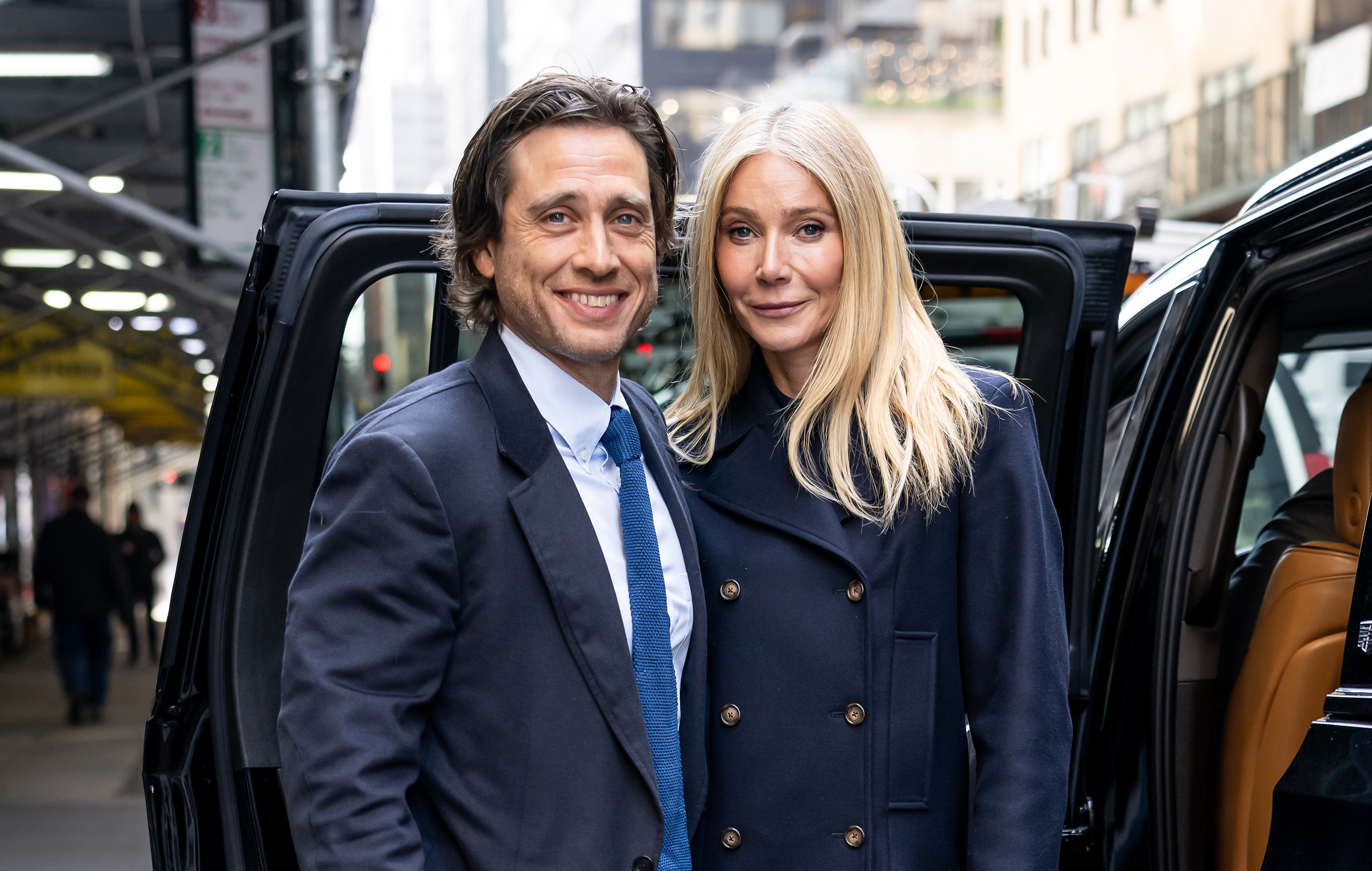 Gwyneth Paltrow stands beside Brad Falchuk near a vehicle in New York City, both facing forward as she maintains a composed expression and he smiles toward the camera.