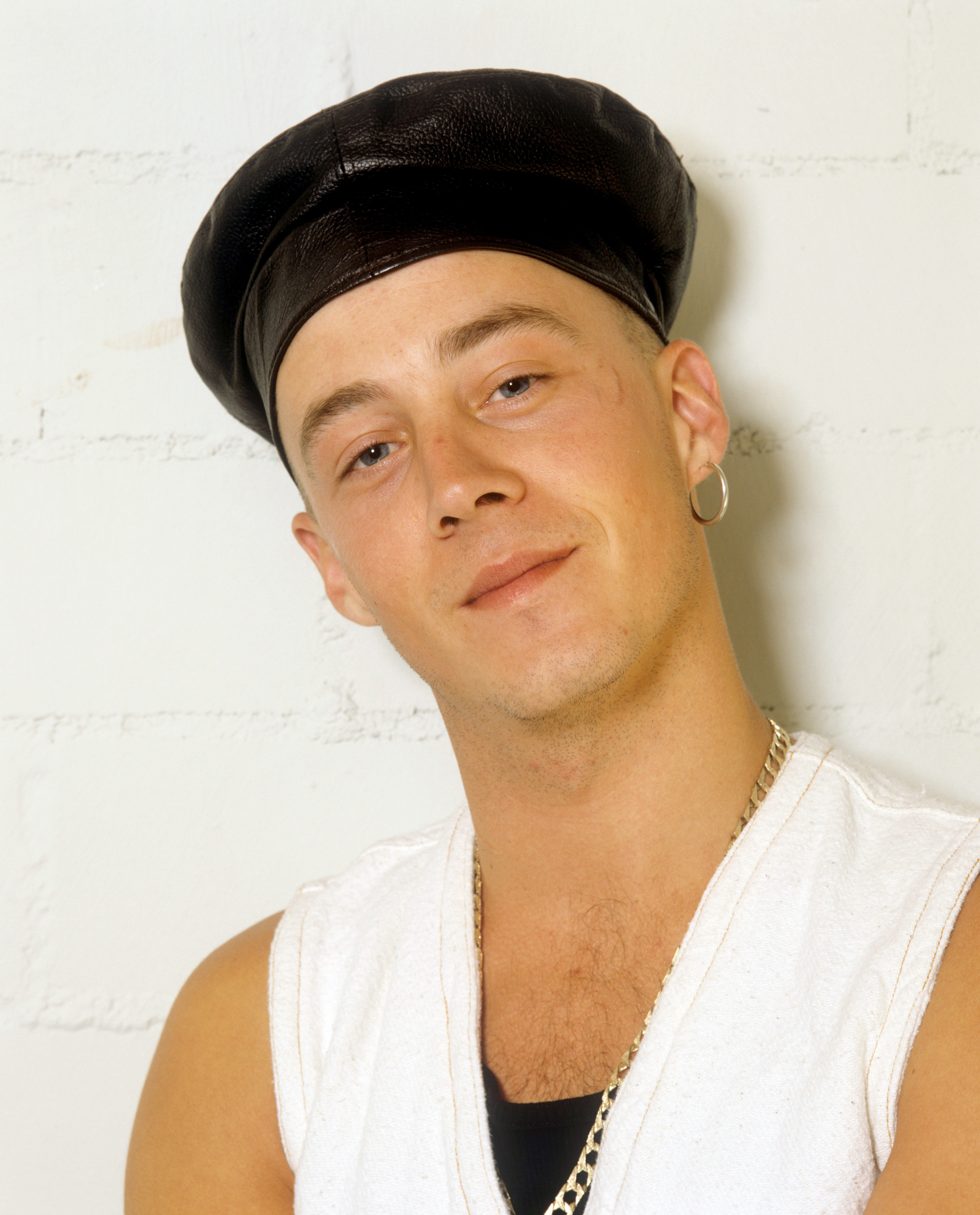 In this close-up taken on 26 June 1993 in Schwenningen, Germany, John Hendy leans casually against a wall, his relaxed expression and subtle smile offering a softer contrast to the band's typically bold image. Dressed in a sleeveless top with a leather cap and simple jewellery, the portrait captures a quieter, more personal moment during East 17's early days in the spotlight.