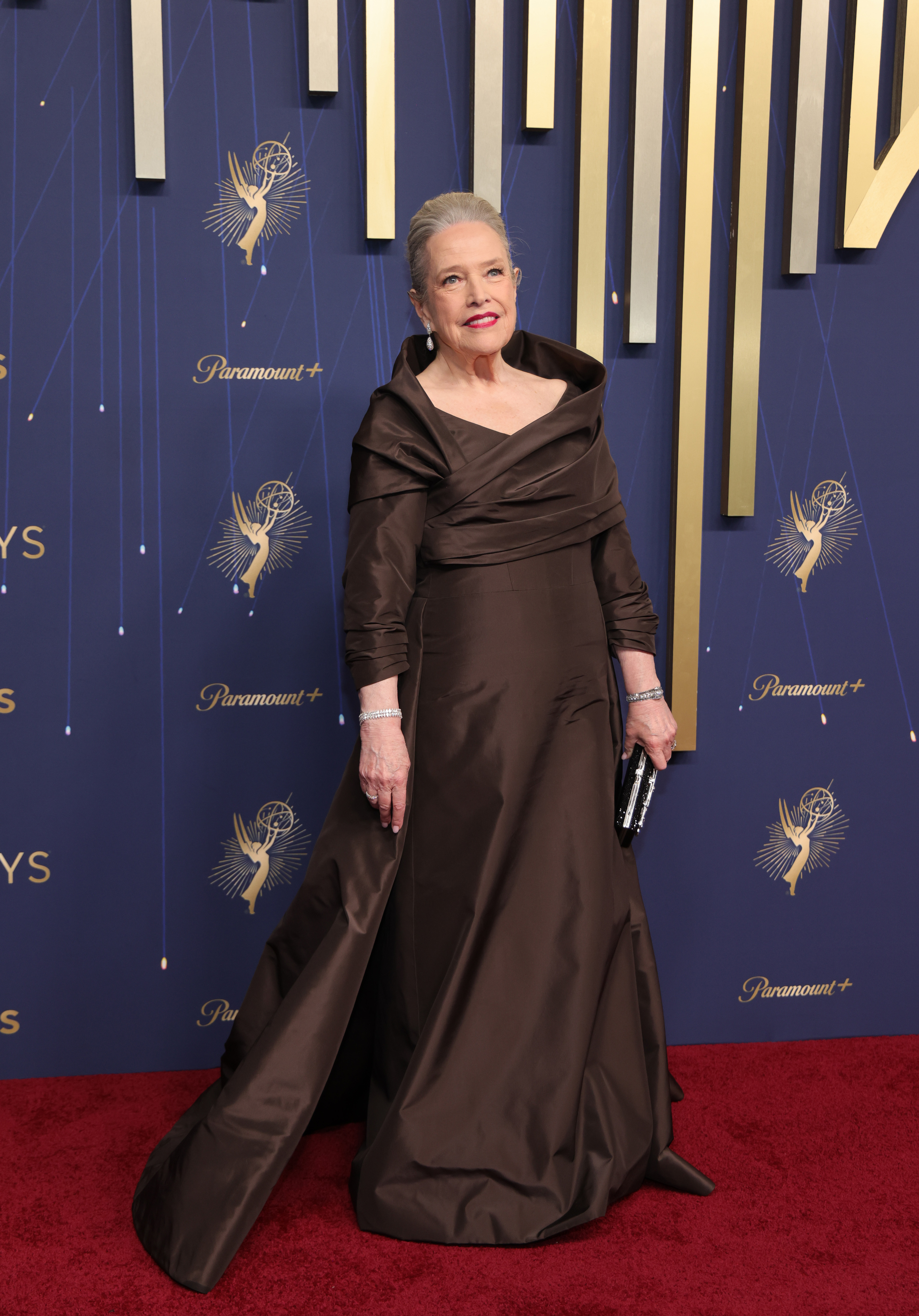 Kathy Bates wore a refined dark floor-length gown with an open neckline to the Emmys. She styled the look with a voluminous updo, a bold red lip, and white gold diamond jewelry, including a double tennis bracelet with pear-cut stones.