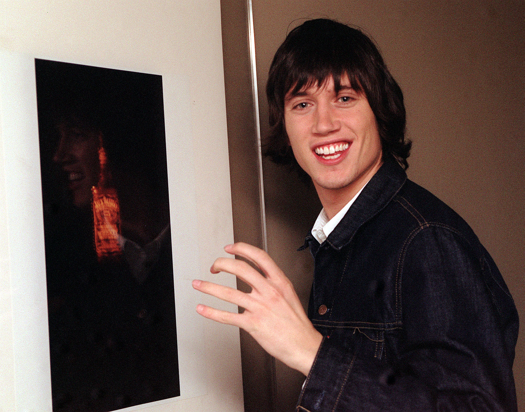 Vernon Kay with shaggy dark hair wears a dark denim jacket over a light shirt, smiling broadly as he gestures toward a framed image on the wall.