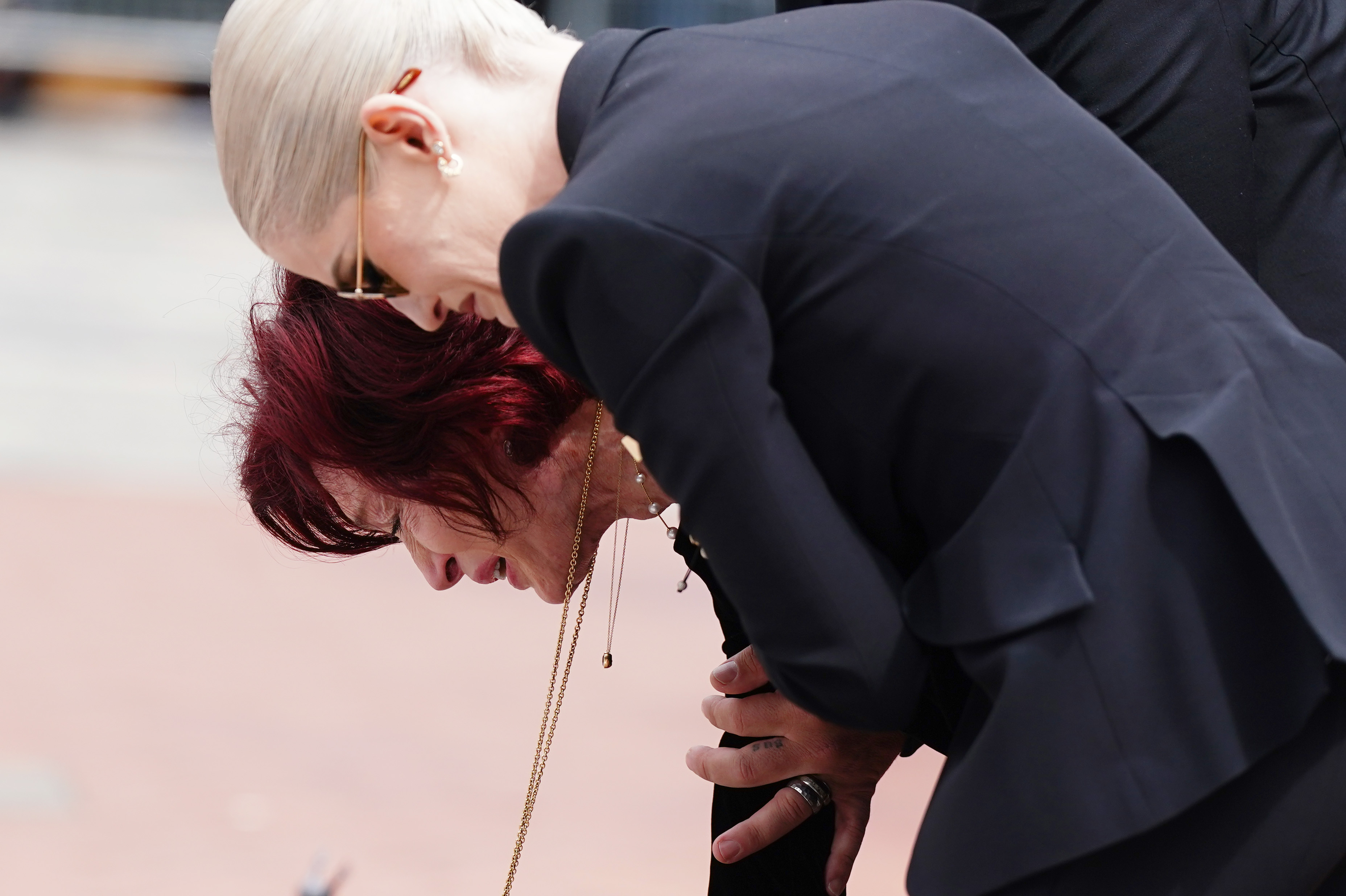 Sharon Osbourne bends forward in visible anguish as her daughter Kelly gently supports her during an emotional moment. Their closeness captures the raw pain and strength shared in mourning at Ozzy's funeral.