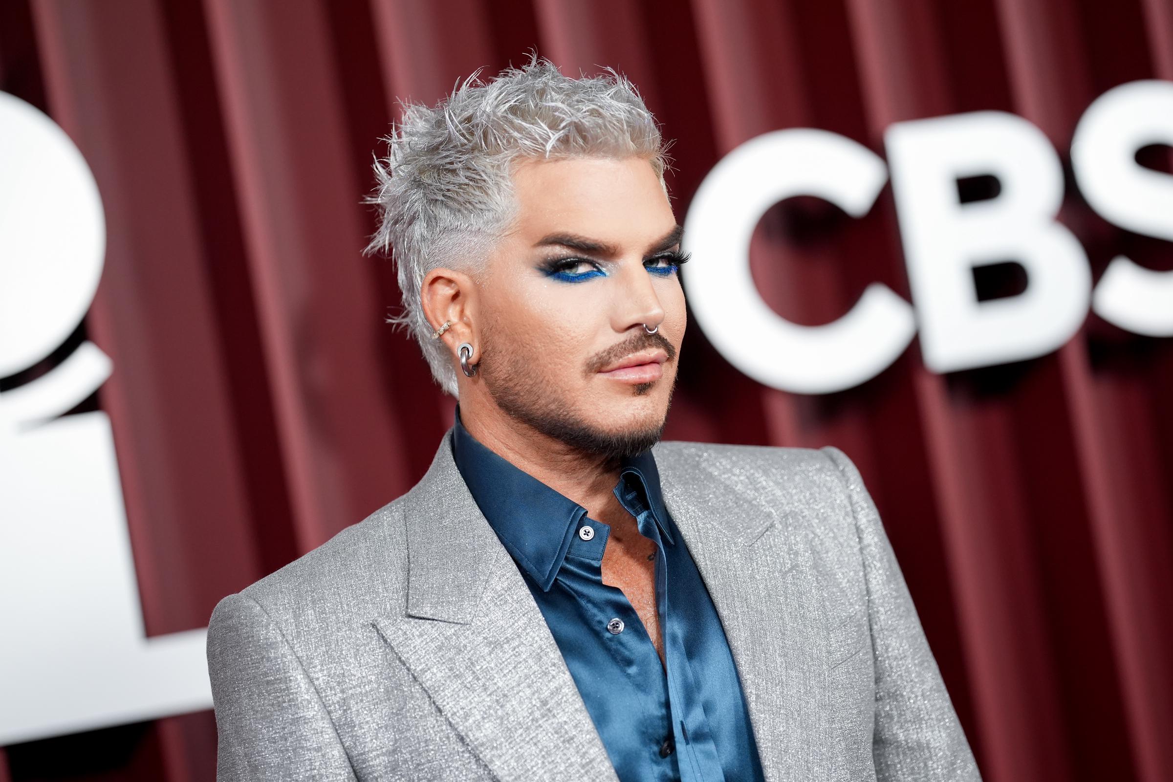 Adam Lambert glances to the side, his soft smirk enhanced by precise contouring and icy hair. The subtle shimmer of his jacket and the depth of his eye makeup form a look that’s sleek, modern, and unmistakably infused with star quality.