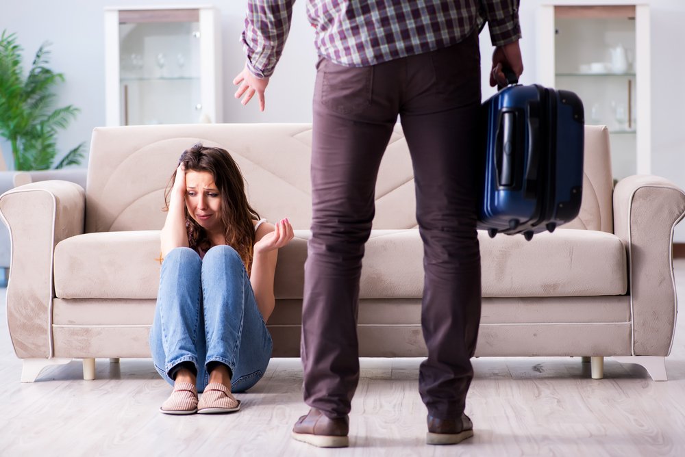 A woman sits crying against a couch while her man stands in front of her holding a travel bag | Photo: Shutterstock/Elnur