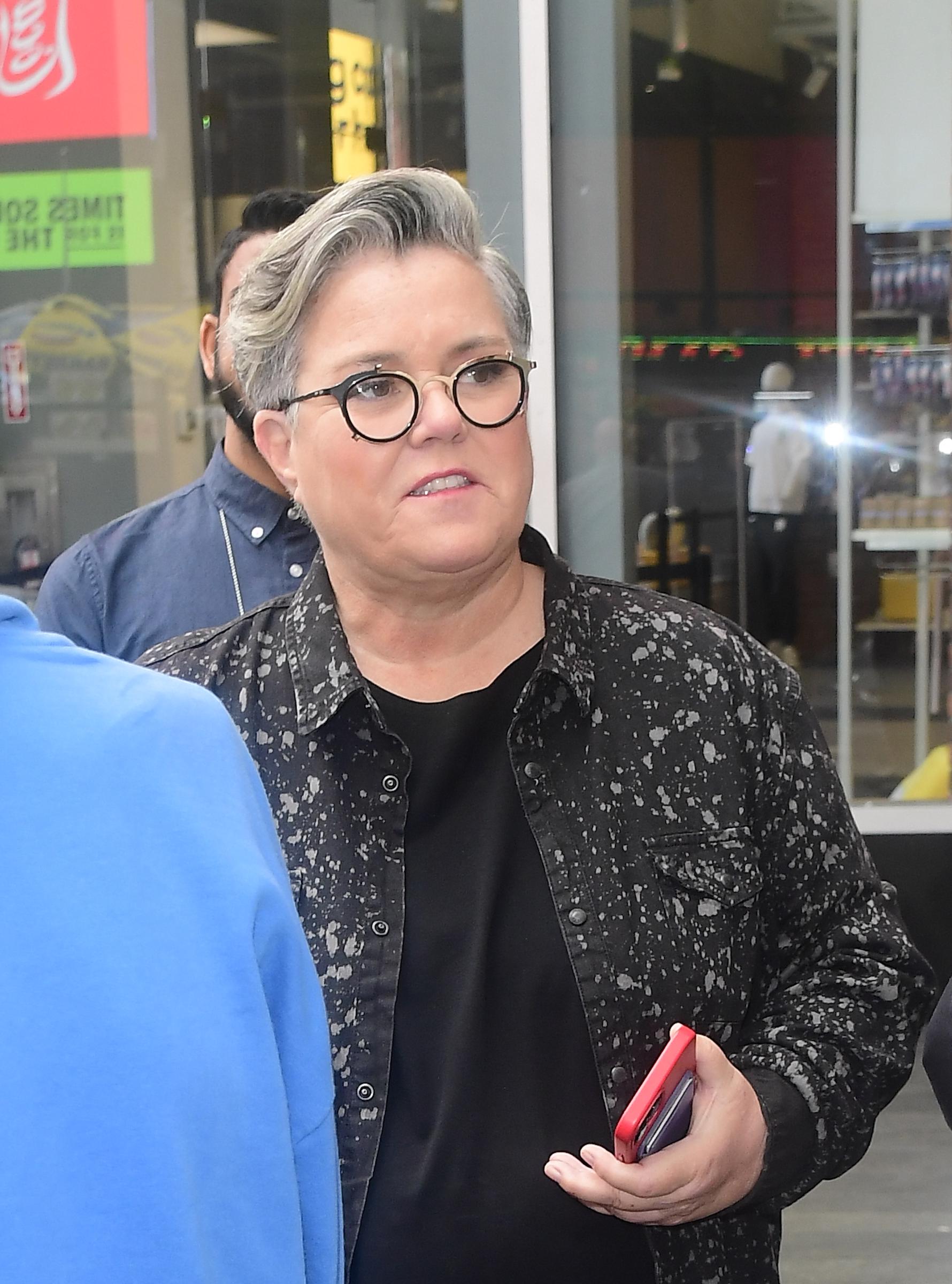 Rosie O'Donnell is photographed during a casual outing in Midtown Manhattan, wearing a black patterned jacket and round glasses while holding a phone on a busy street.
