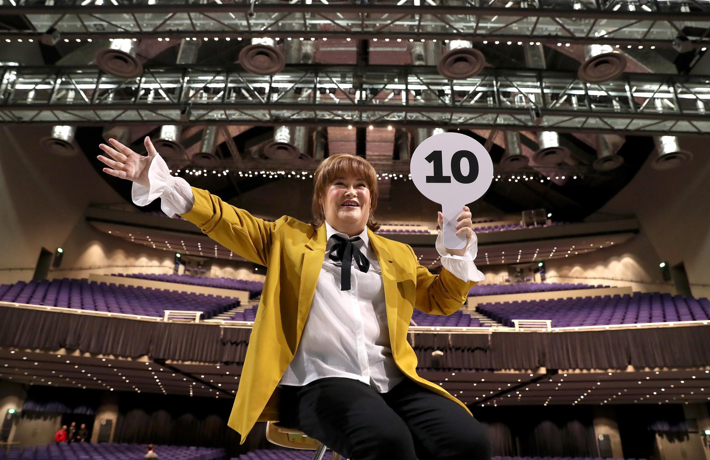 Boyle perches on a chair at the empty SEC Armadillo in Glasgow on 18 February 2020, one arm flung wide and the other holding up a scorecard reading "10," as she promotes her "Ten Tour" at the very venue where she famously auditioned a decade earlier.