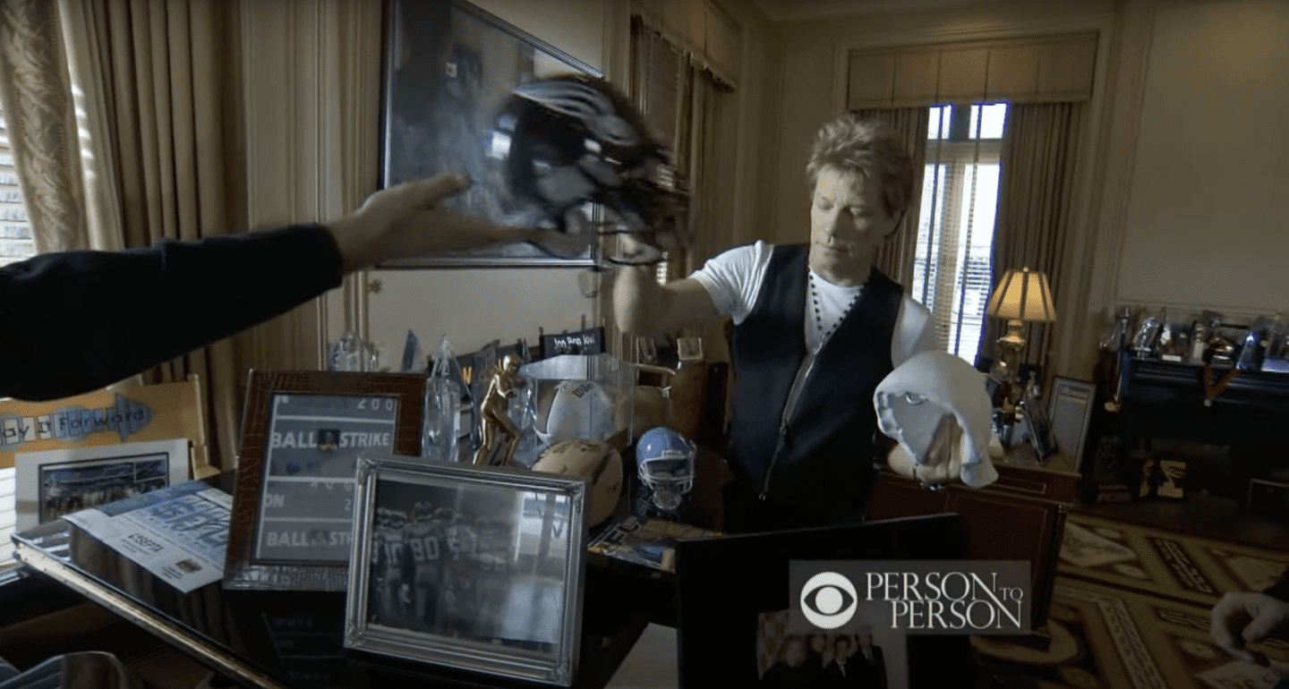 Jon Bon Jovi pictured holding helmets in his home office where he keeps all his gifts and awards. / Source: YouTube/@CBS