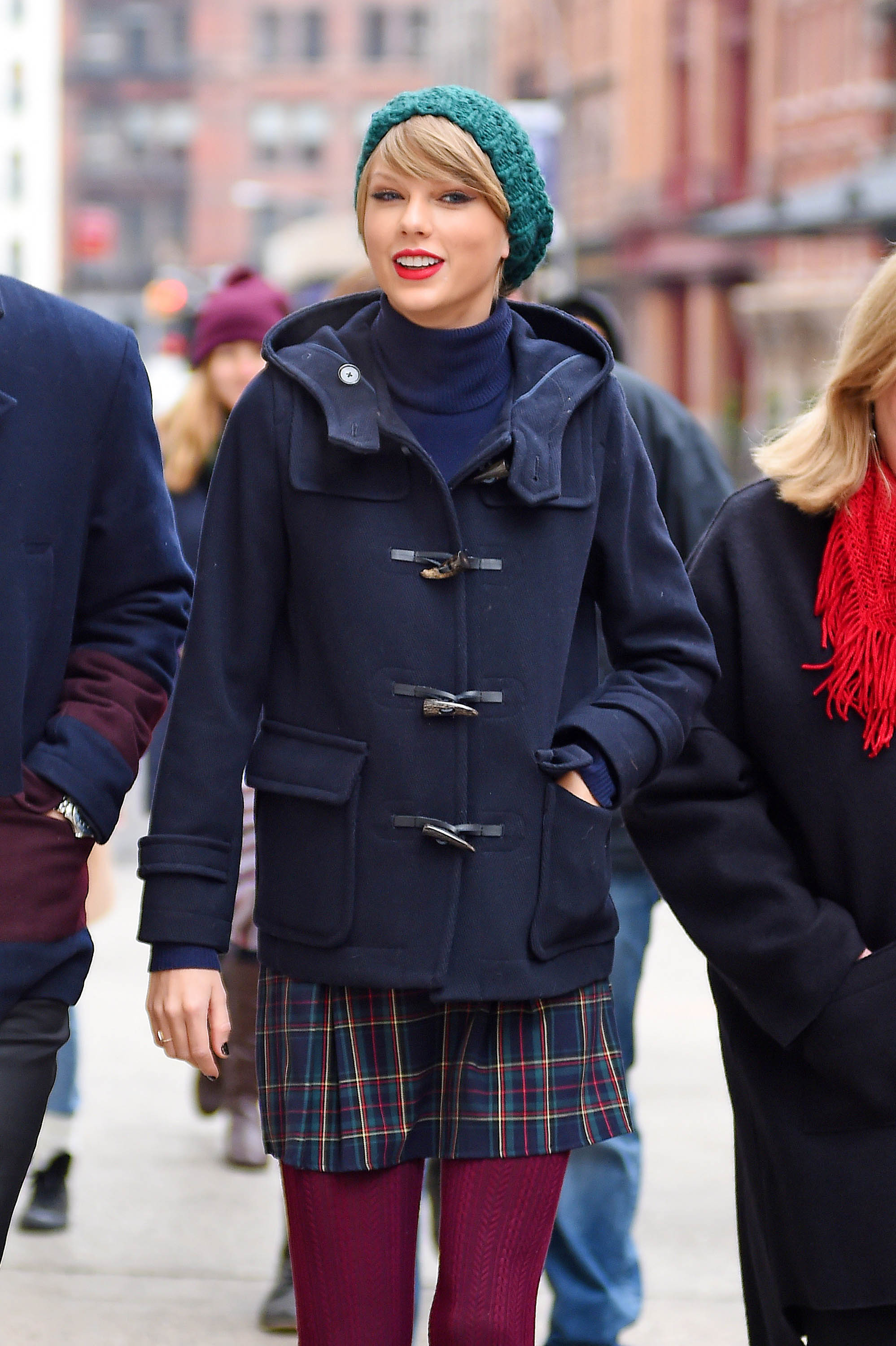 Singer Taylor Swift is spotted in New York City wearing a navy duffle coat over a plaid skirt with burgundy tights and a teal knit beanie. Her bright red lipstick adds a vibrant pop to the chic and cozy winter street style.