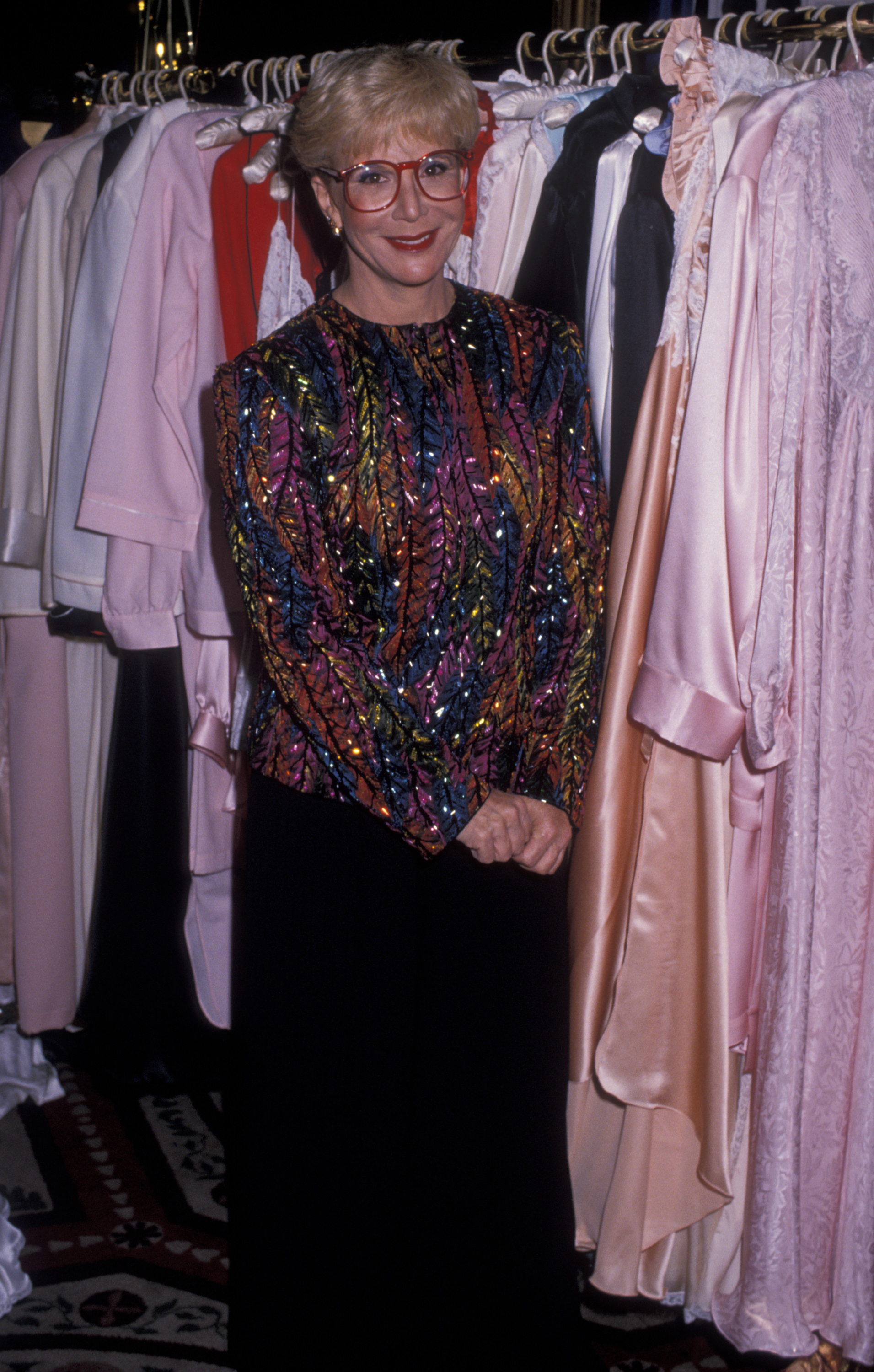 Sally Jessy Raphael poses among elegant garments at The Promise Ball benefiting the Juvenile Diabetes Foundation, held at the Waldorf Hotel in New York City on November 11, 1989. Her sequined, multicolored top and signature red glasses add sparkle to the charitable evening.