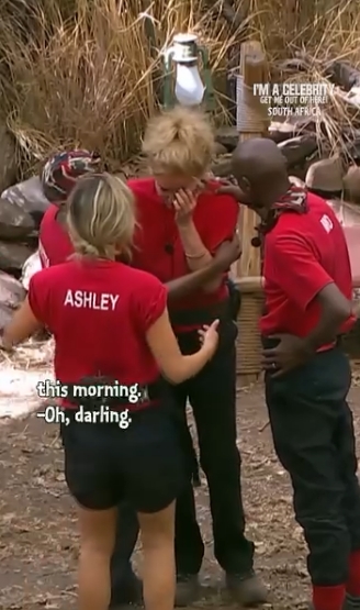 Standing in the camp clearing, Beverley Callard appears visibly shaken as she breaks the news to her fellow contestants, with Ashley Banjo stepping closer in concern while others gather around to comfort her during the difficult announcement. | Source: Instagram/imacelebrity