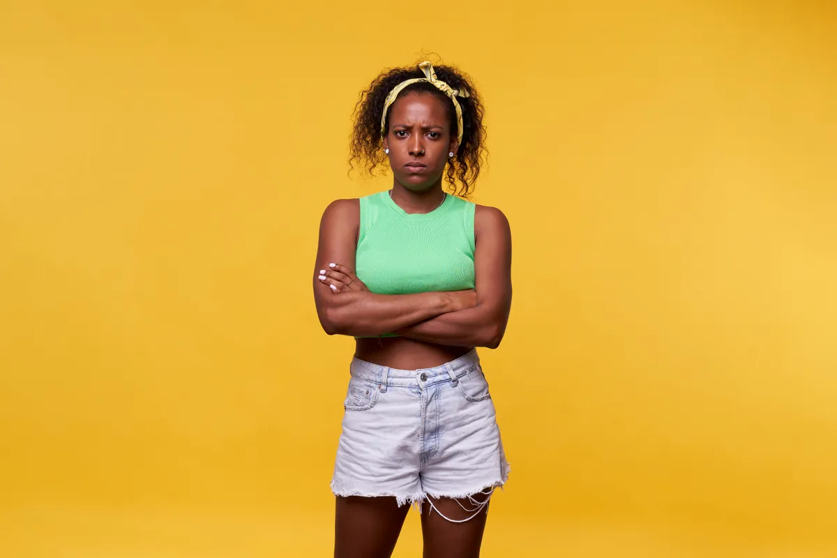 An angry Latin woman standing against a yellow background crossing her arms and looking at the camera seriously. | Source: Getty Images