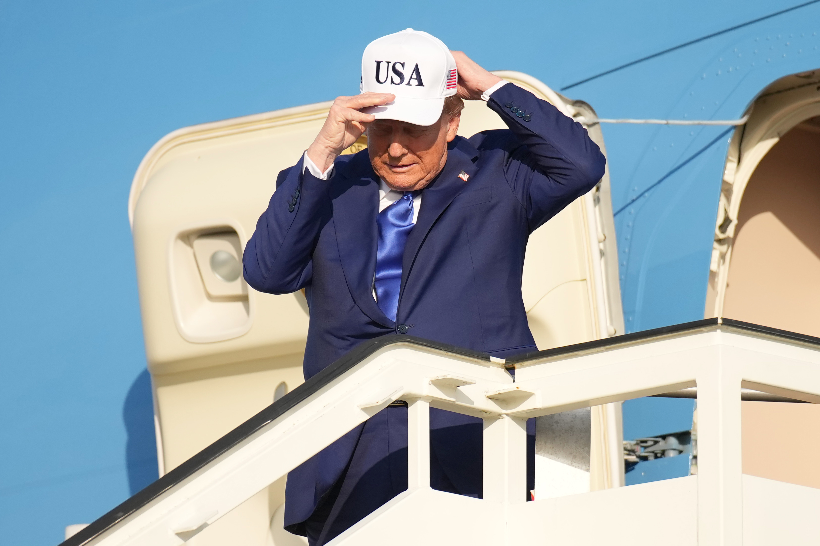 Trump is seen at the top of the airplane stairs at Amsterdam Airport Schiphol, wearing a white cap with “USA” and an American flag printed on it as he arrives ahead of the 2025 NATO Summit.