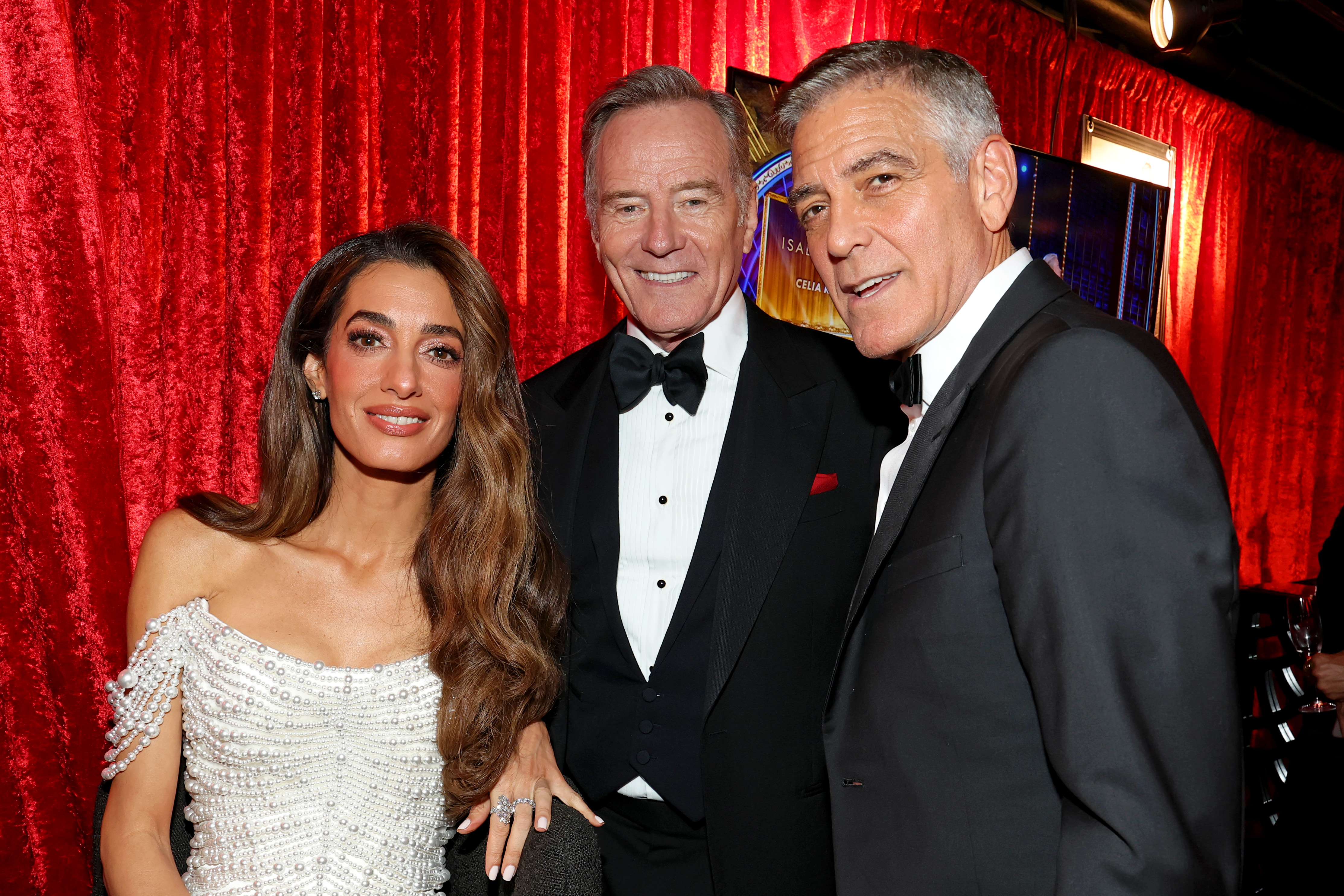 Amal and George Clooney join Bryan Cranston backstage in the Green Room. Framed by red velvet curtains, Amal smiles directly at the camera, while George angles toward her with an easy, relaxed expression.